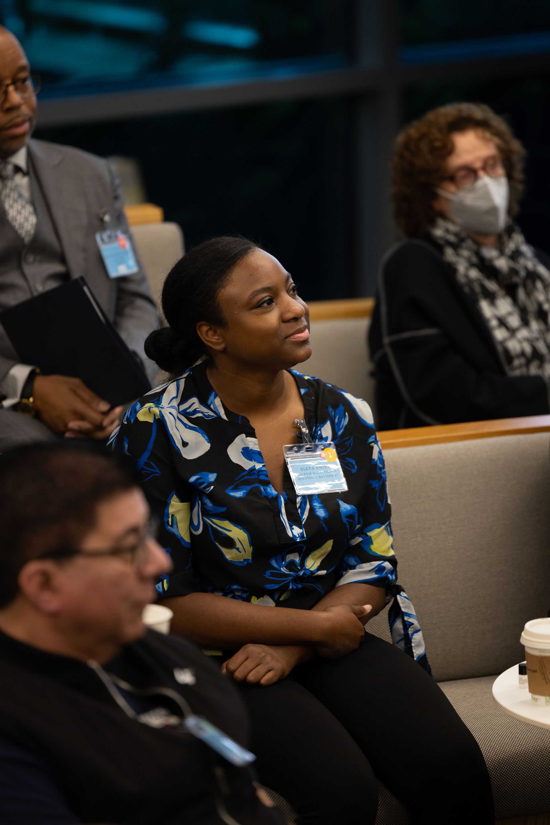 Audience seated in a meeting room, including a woman in a blue floral blouse listening attentively.