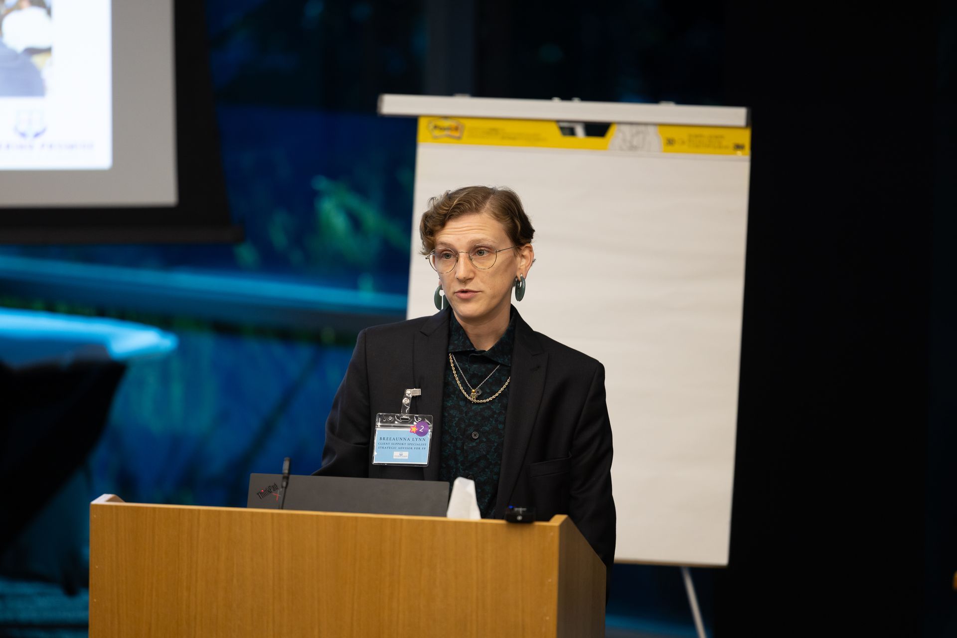 Speaker at a podium in a conference room, with a blank flip chart behind her.