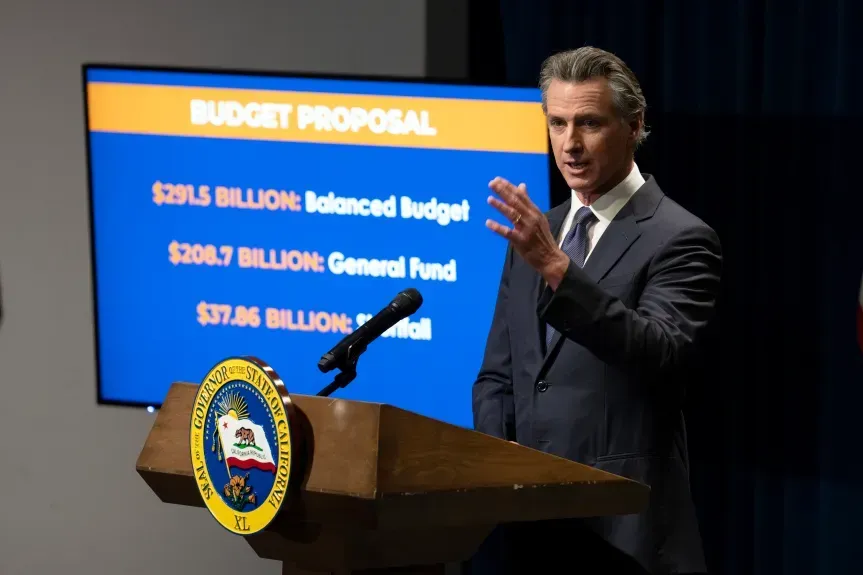 Man speaking at podium beside budget proposal screen in a press briefing room