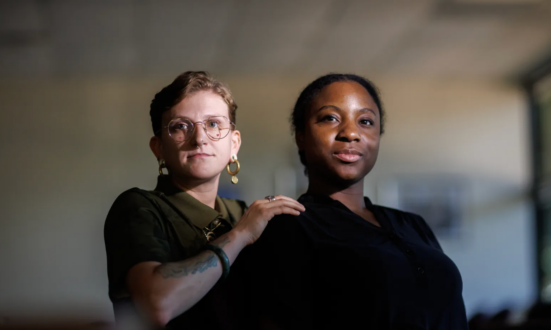 Two people standing together indoors, one resting a hand on the other's shoulder.