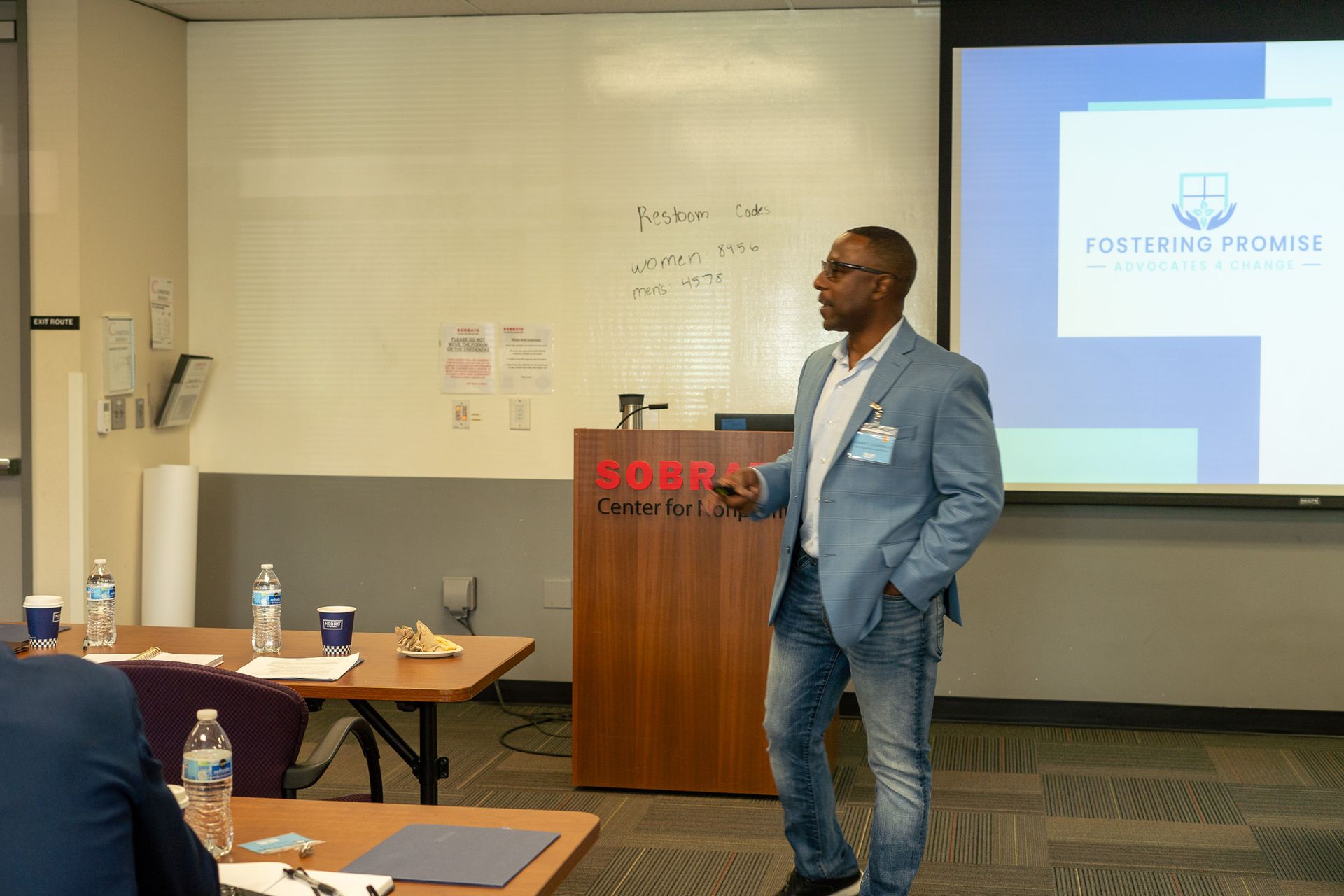 Speaker in blue blazer presenting beside a projector screen in a conference room.
