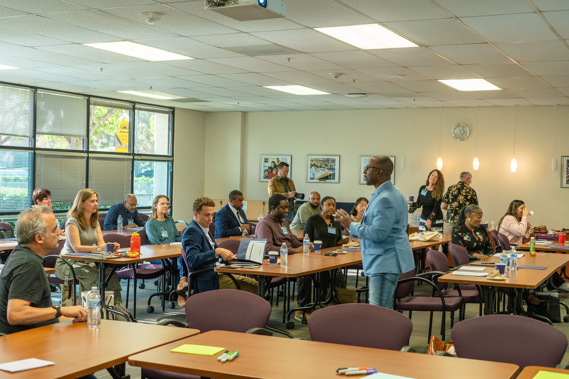Community meeting in a bright room, with people seated at tables and a speaker standing near the front.