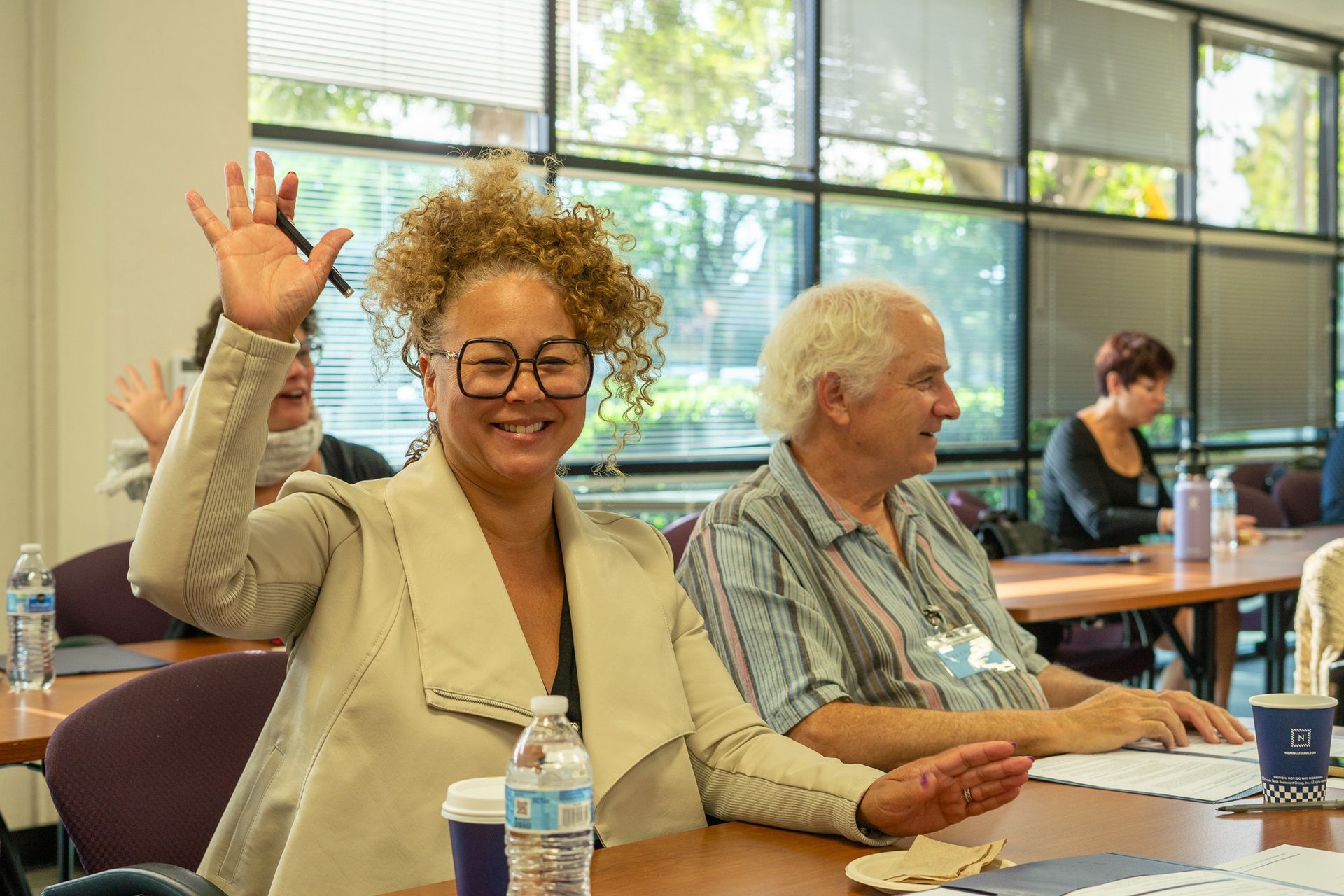 Woman smiling and waving in a bright classroom, seated at a desk with others nearby.