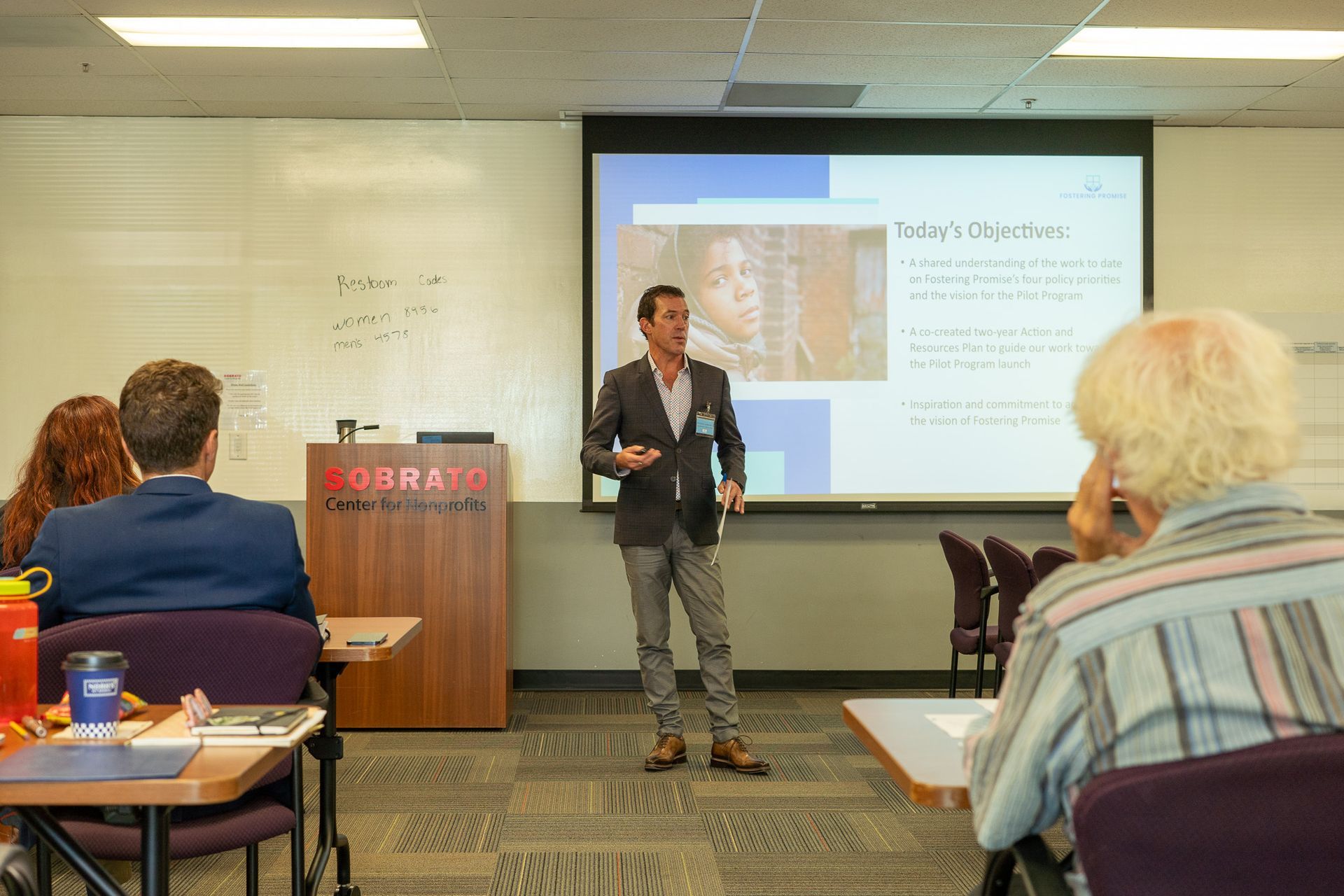 Presenter speaks in a conference room beside a projected slide, with attendees seated and listening