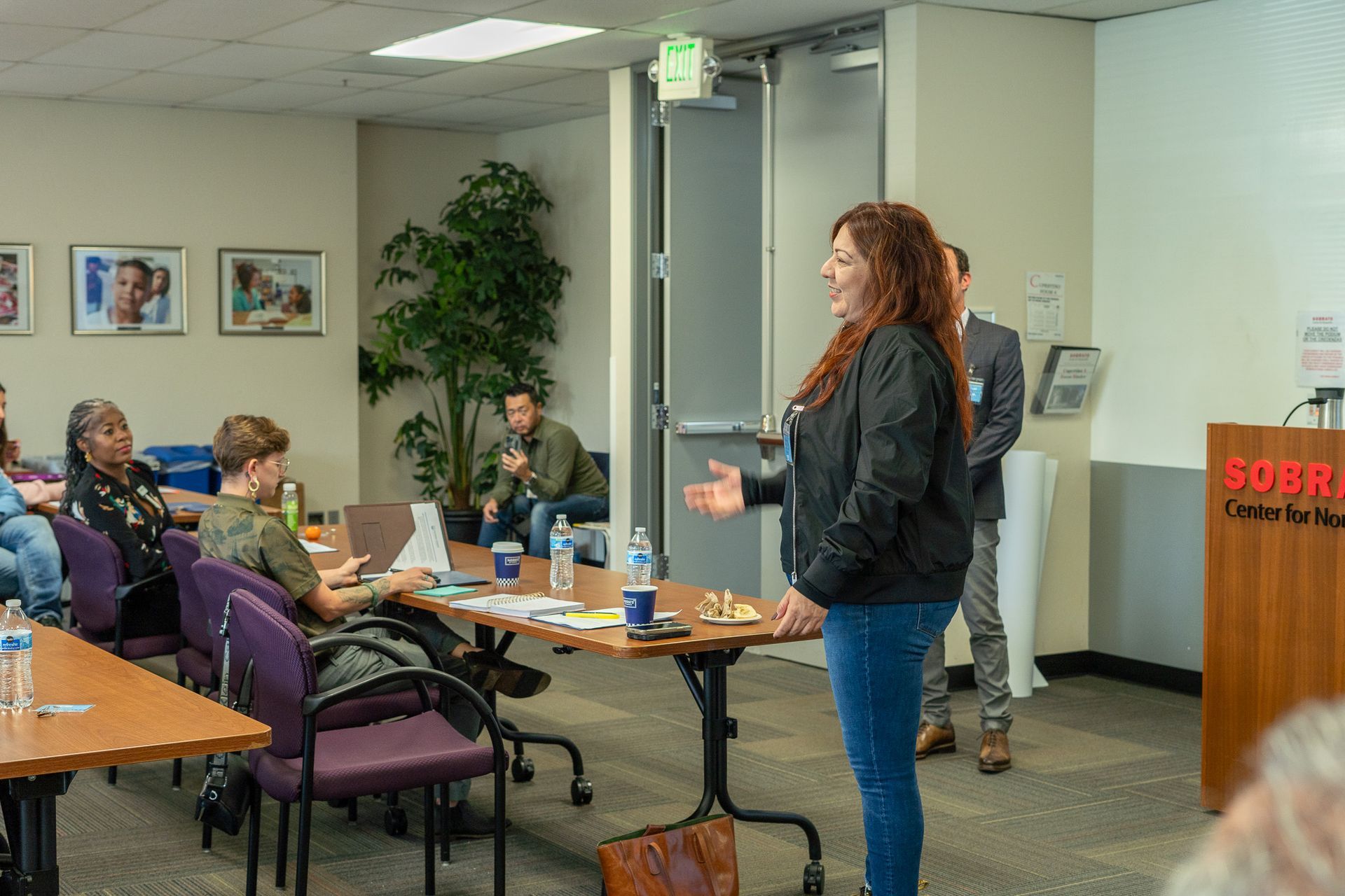Person presenting in a conference room to a seated audience, with tables, chairs, and a podium nearby.