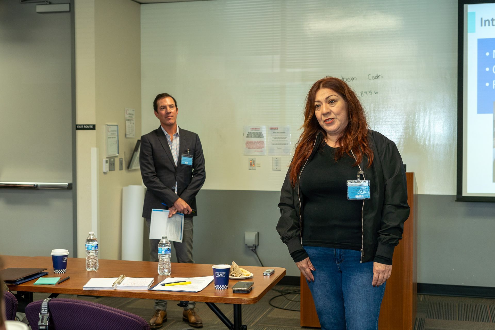 Two people standing in a conference room beside a table with water bottles and papers, presenting to an audience.