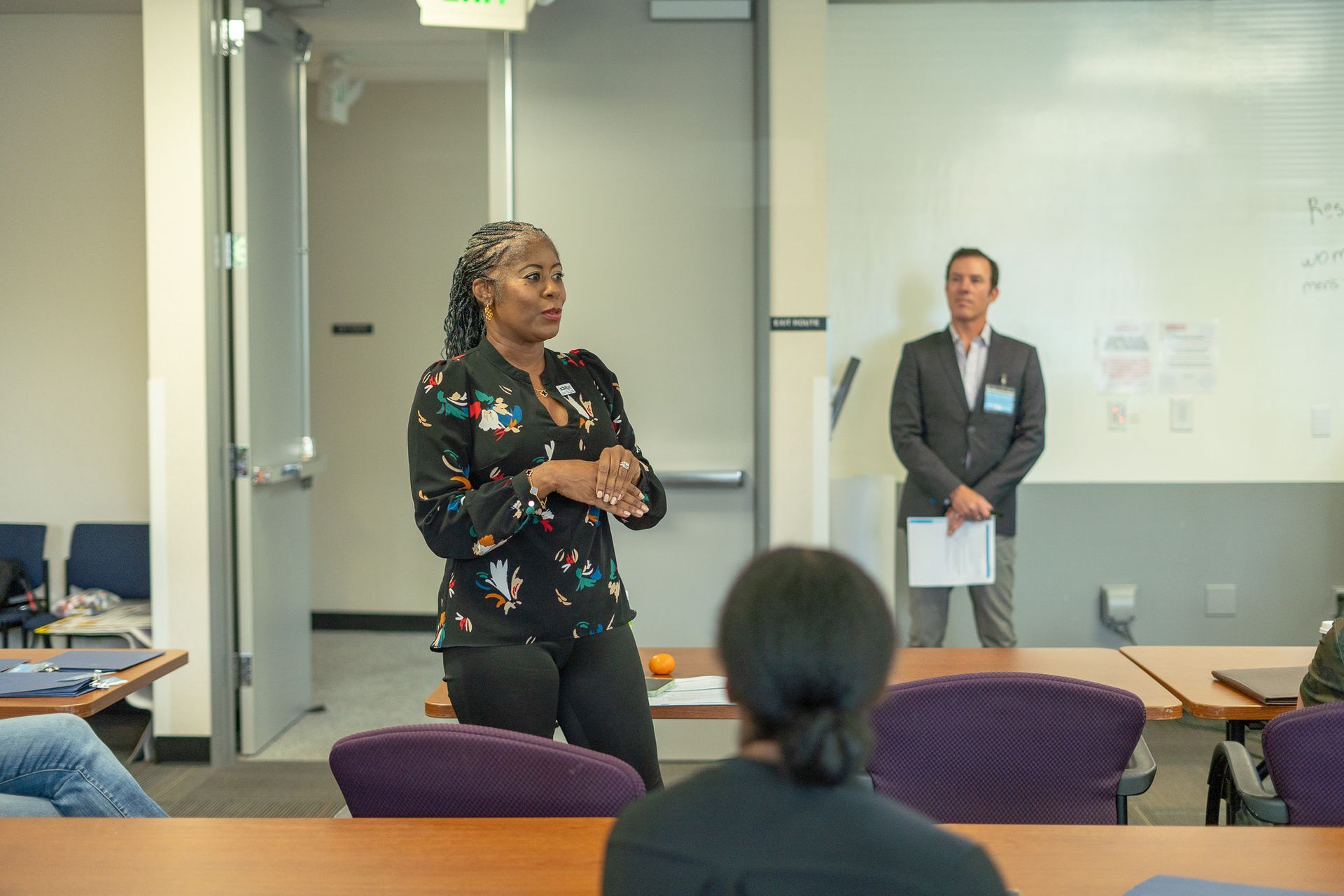 Presenter speaking at the front of a classroom while another person stands nearby, audience seated in purple chairs.