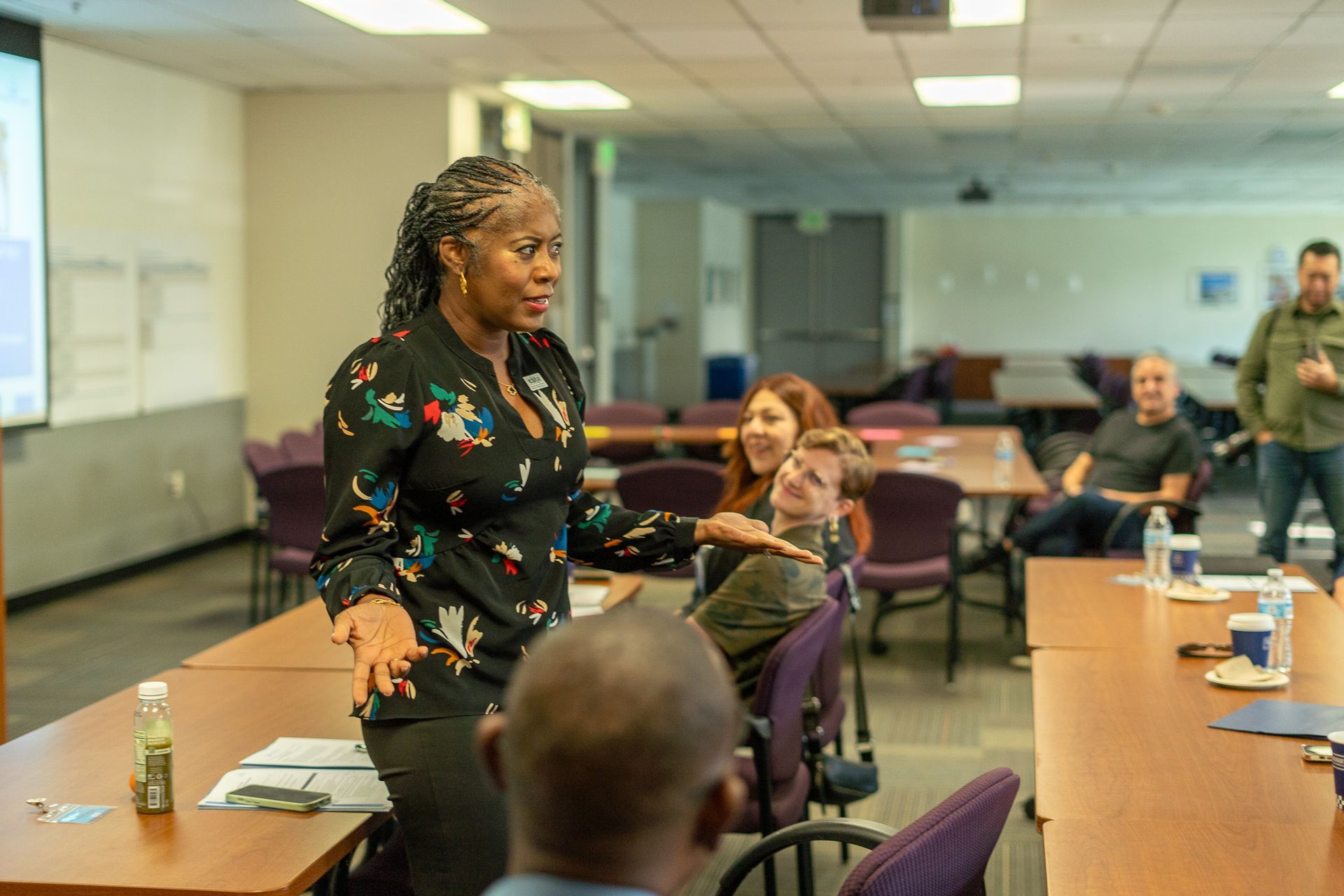 Speaker presenting to a small group in a classroom meeting room, with people seated around tables.
