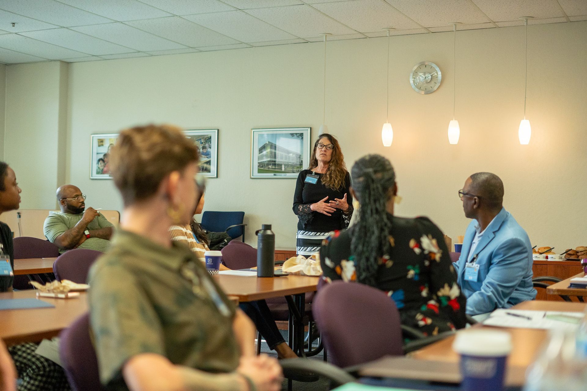 Person speaking at the front of a meeting room while attendees sit around tables listening.