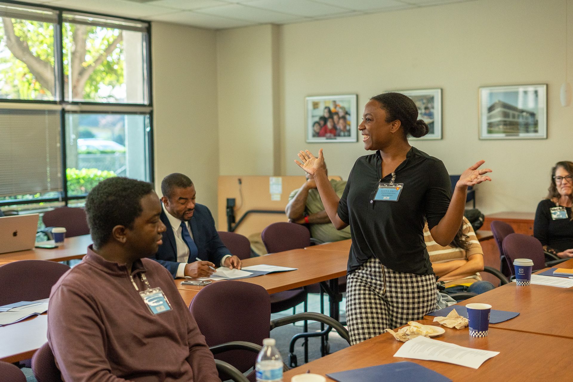 Workshop attendees discuss at tables while a woman stands speaking in a classroom meeting room.