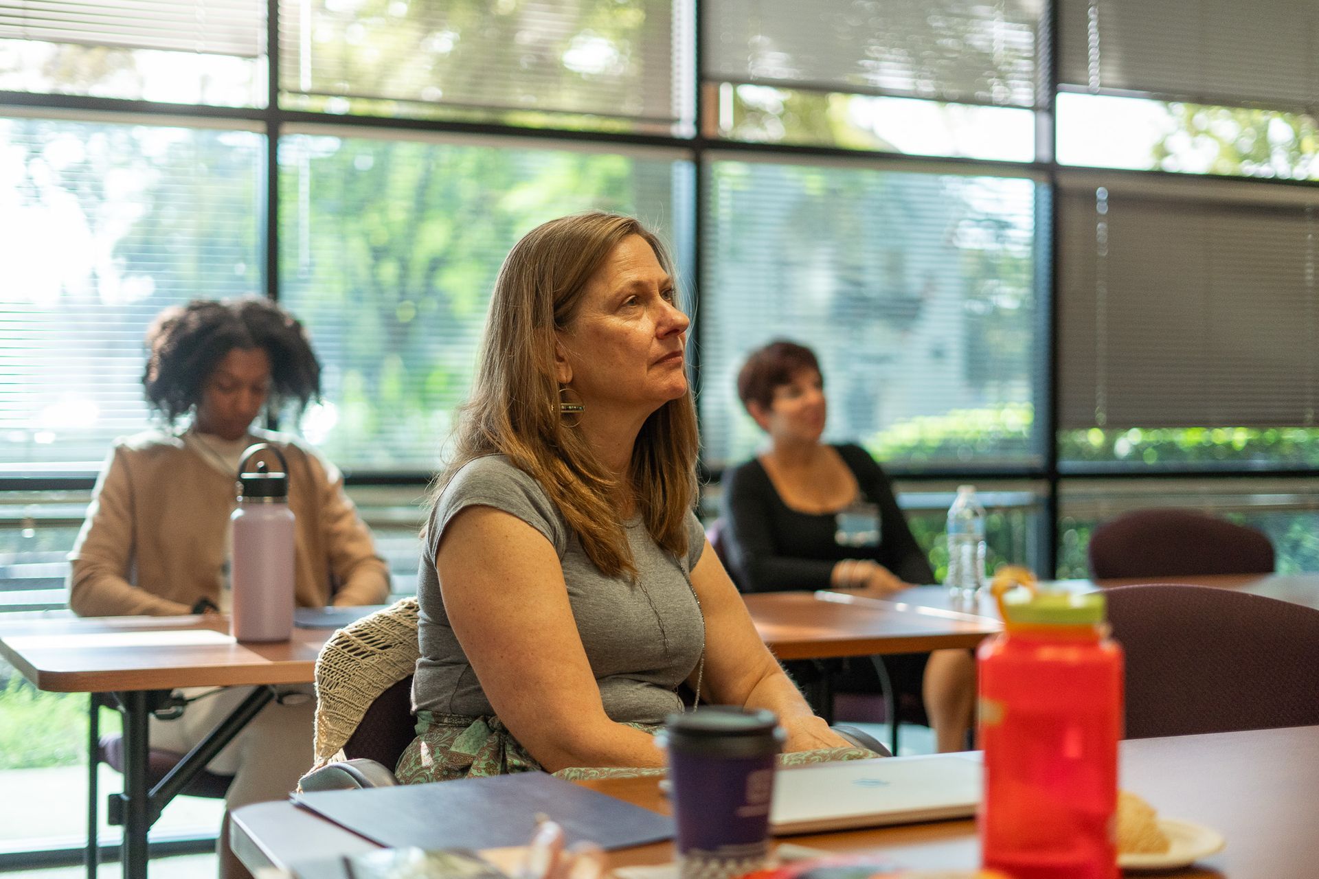 People seated at tables in a bright classroom with large windows and water bottles