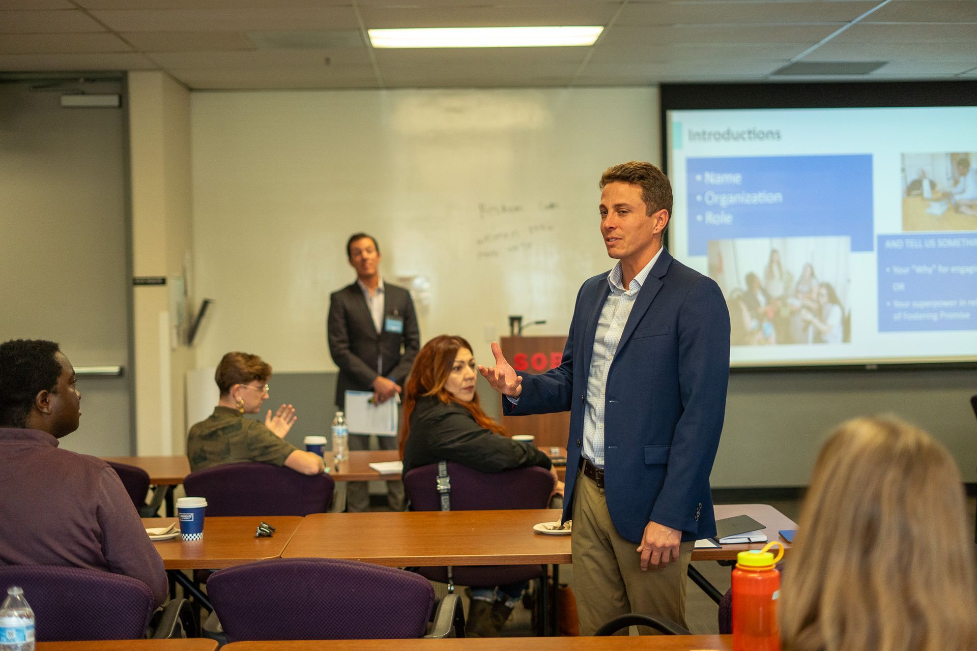 Man presenting in a classroom to seated attendees, with a projected slide on the wall.