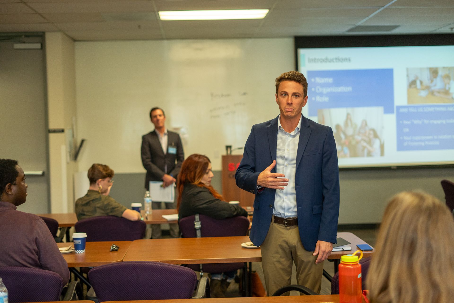 Presenter speaking in a classroom while attendees sit at tables and watch a projected slide.