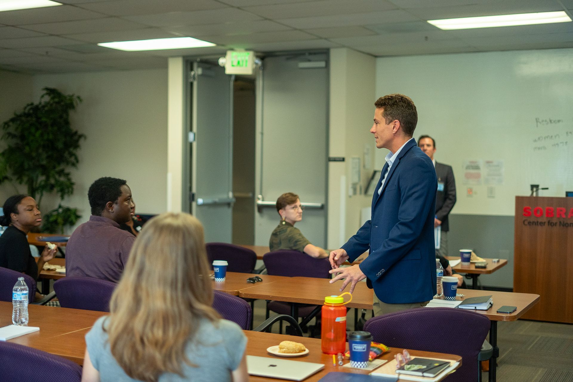Presenter speaking to seated audience in a conference room, with a whiteboard and water bottles on the table.