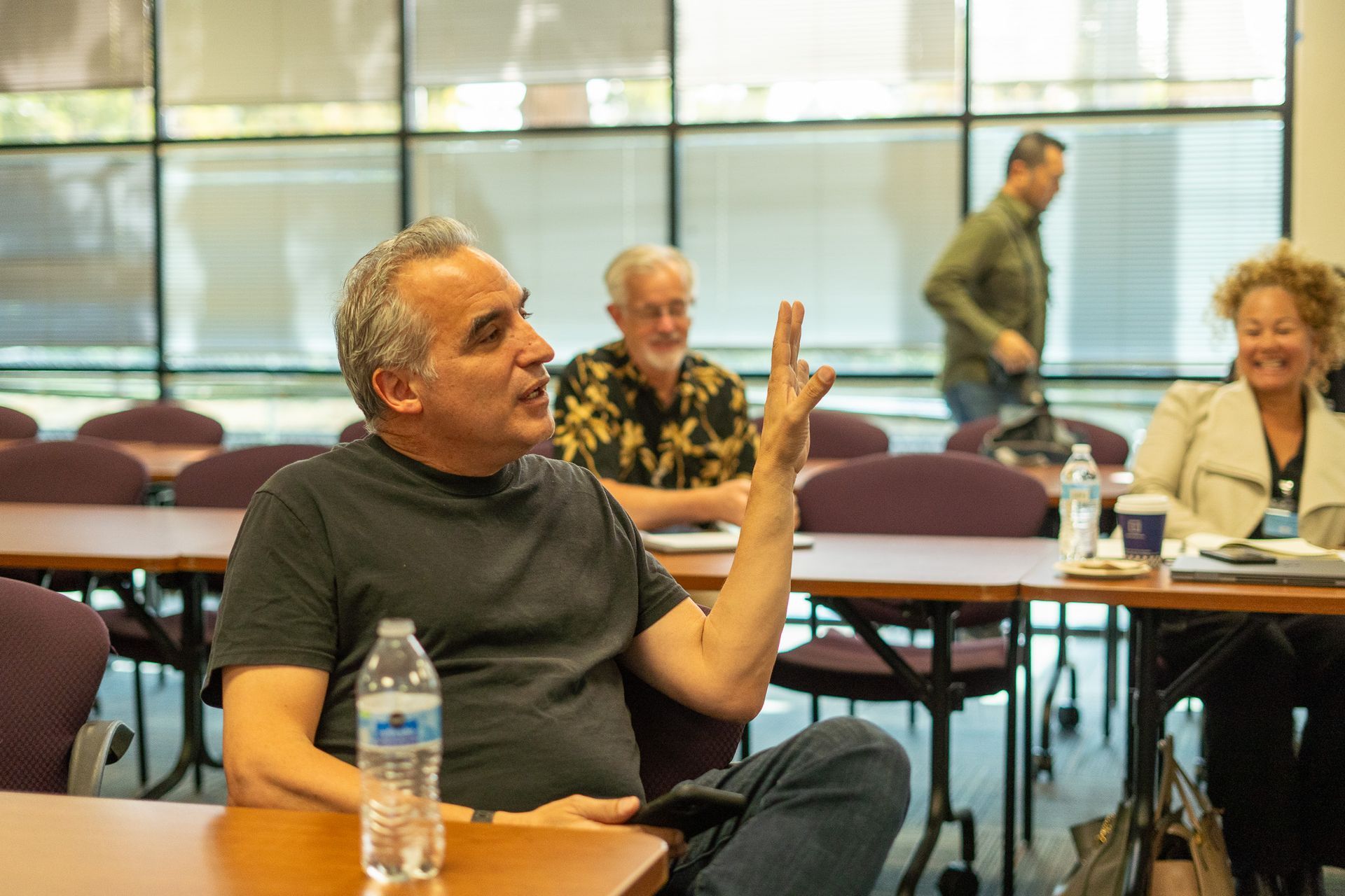 Man speaking and gesturing in a meeting room, with seated attendees and tables in the background.