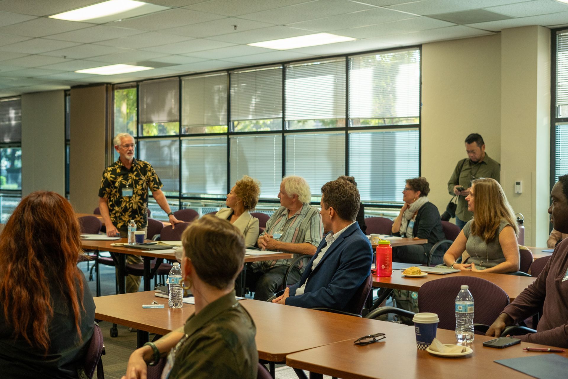 People seated in a classroom listening to a speaker standing near the front windows