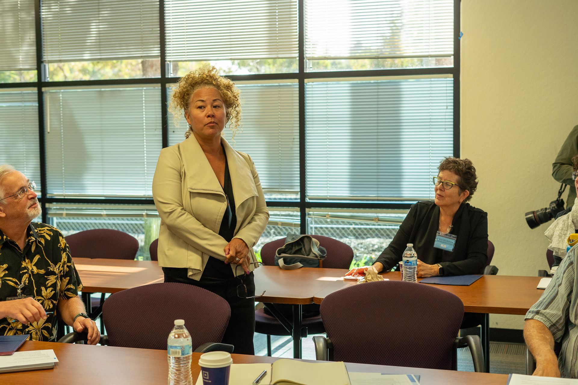 People seated in a meeting room while a woman stands speaking near tables and large windows