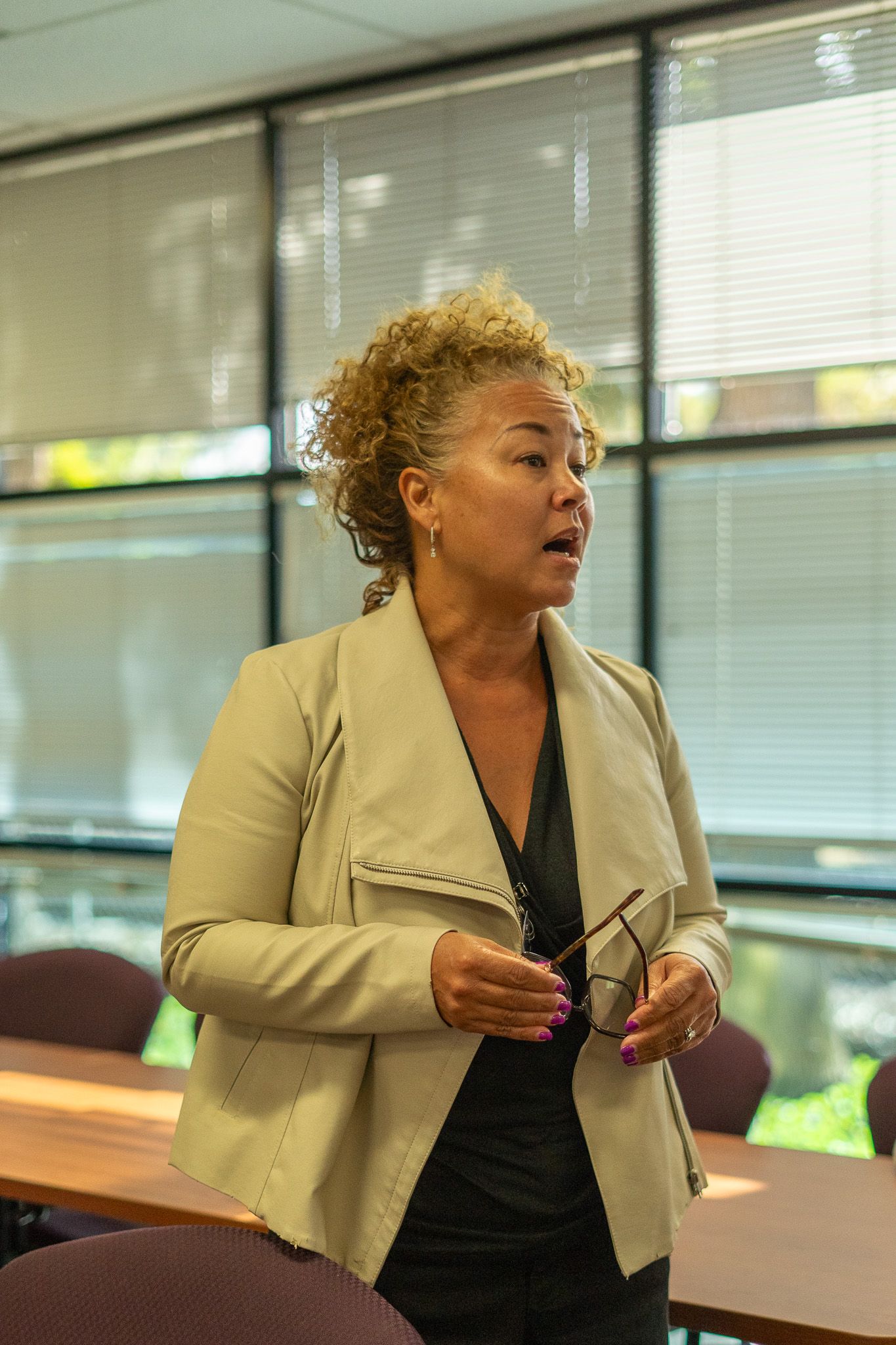 Woman speaking in a conference room, holding glasses near a table and windows.