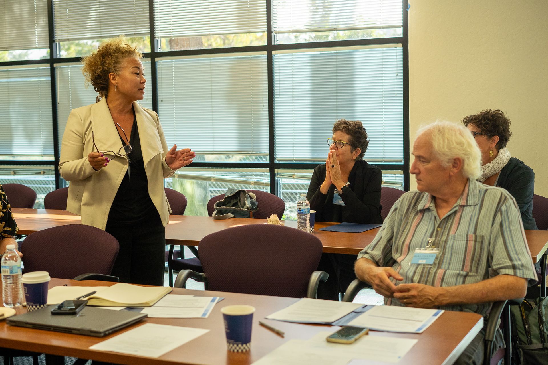 Workshop attendees seated at tables while a woman speaks, in a bright meeting room with large windows.