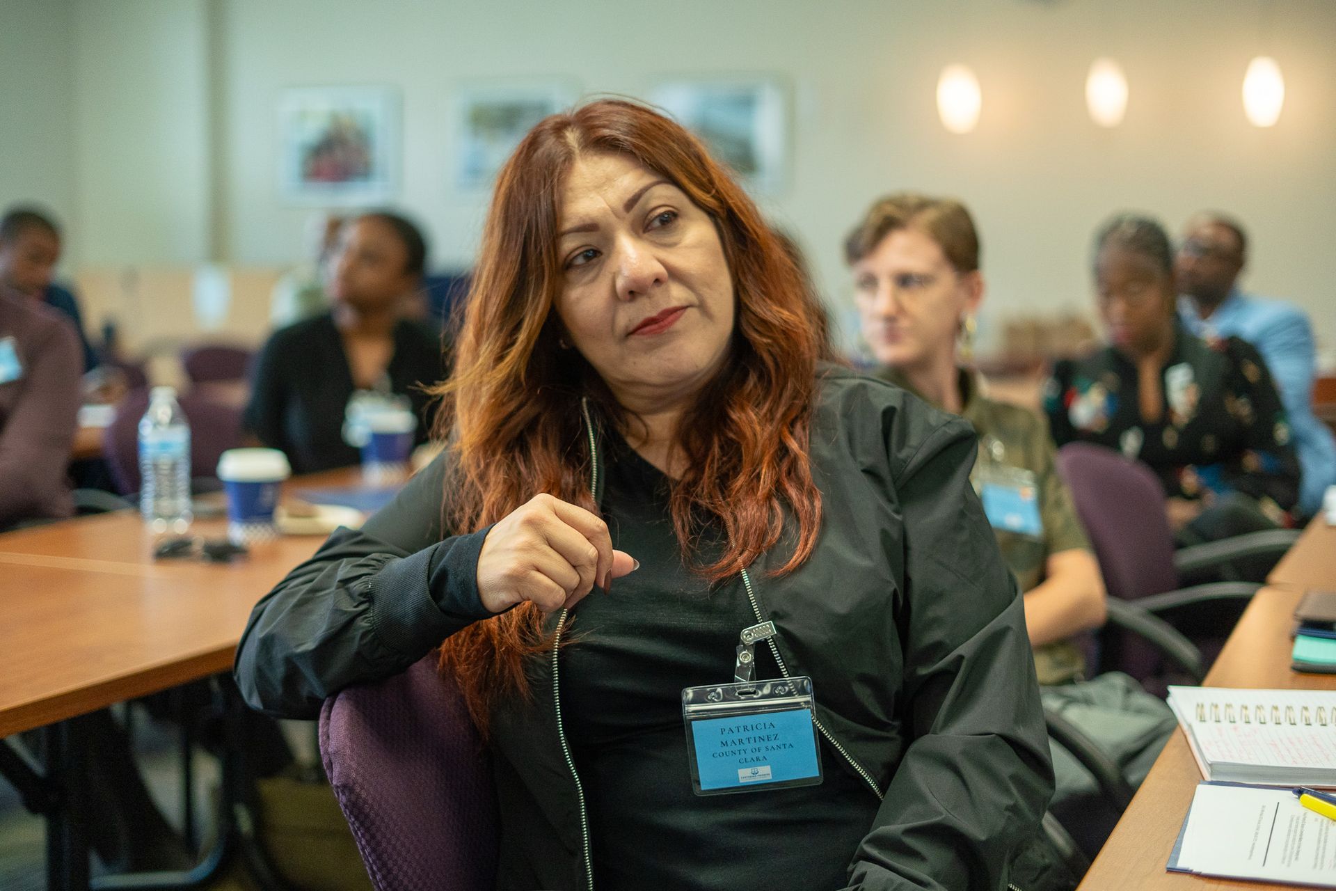 Woman speaking at a conference table, wearing a dark jacket and name badge, with attendees blurred in the background