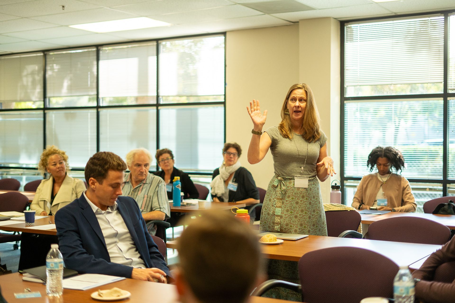 Woman speaking at a conference table while attendees listen in a bright meeting room.