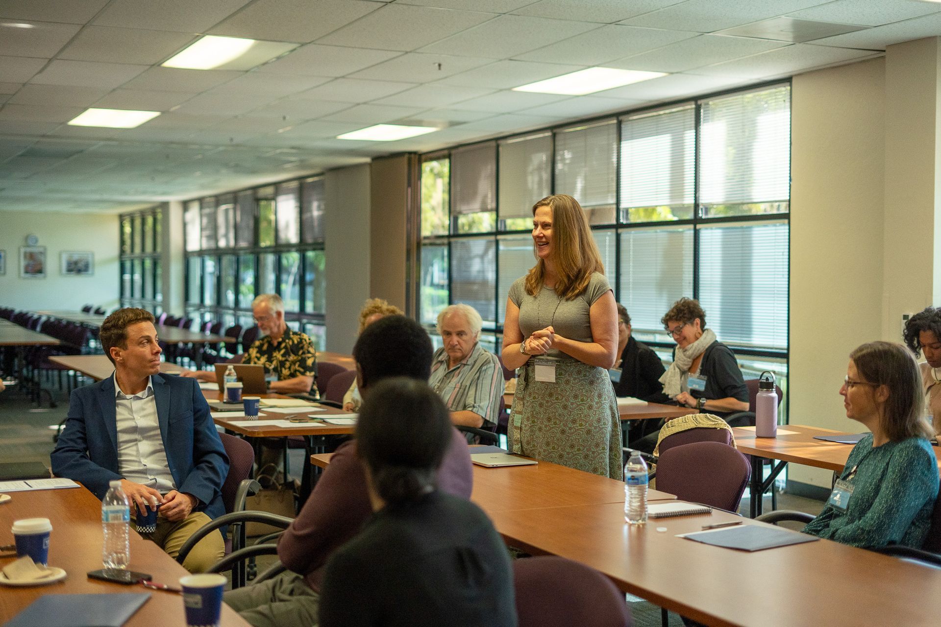 Conference room meeting with people seated at tables, one woman standing and speaking near the front.