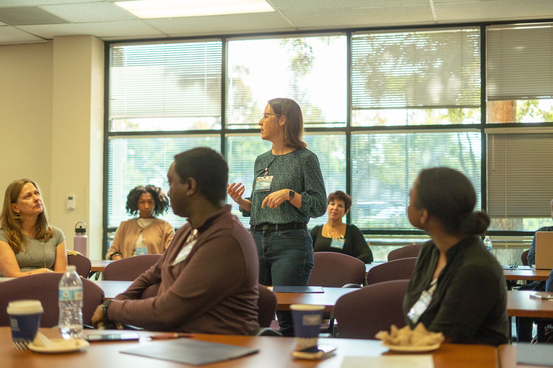 Meeting in a conference room, with one woman standing and speaking while others sit and listen.