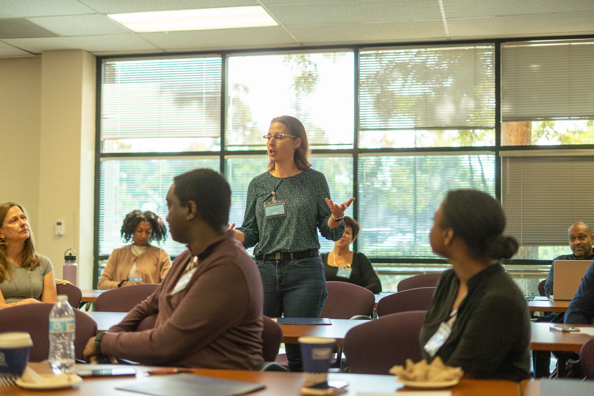 People seated in a meeting room while a woman stands speaking near the windows