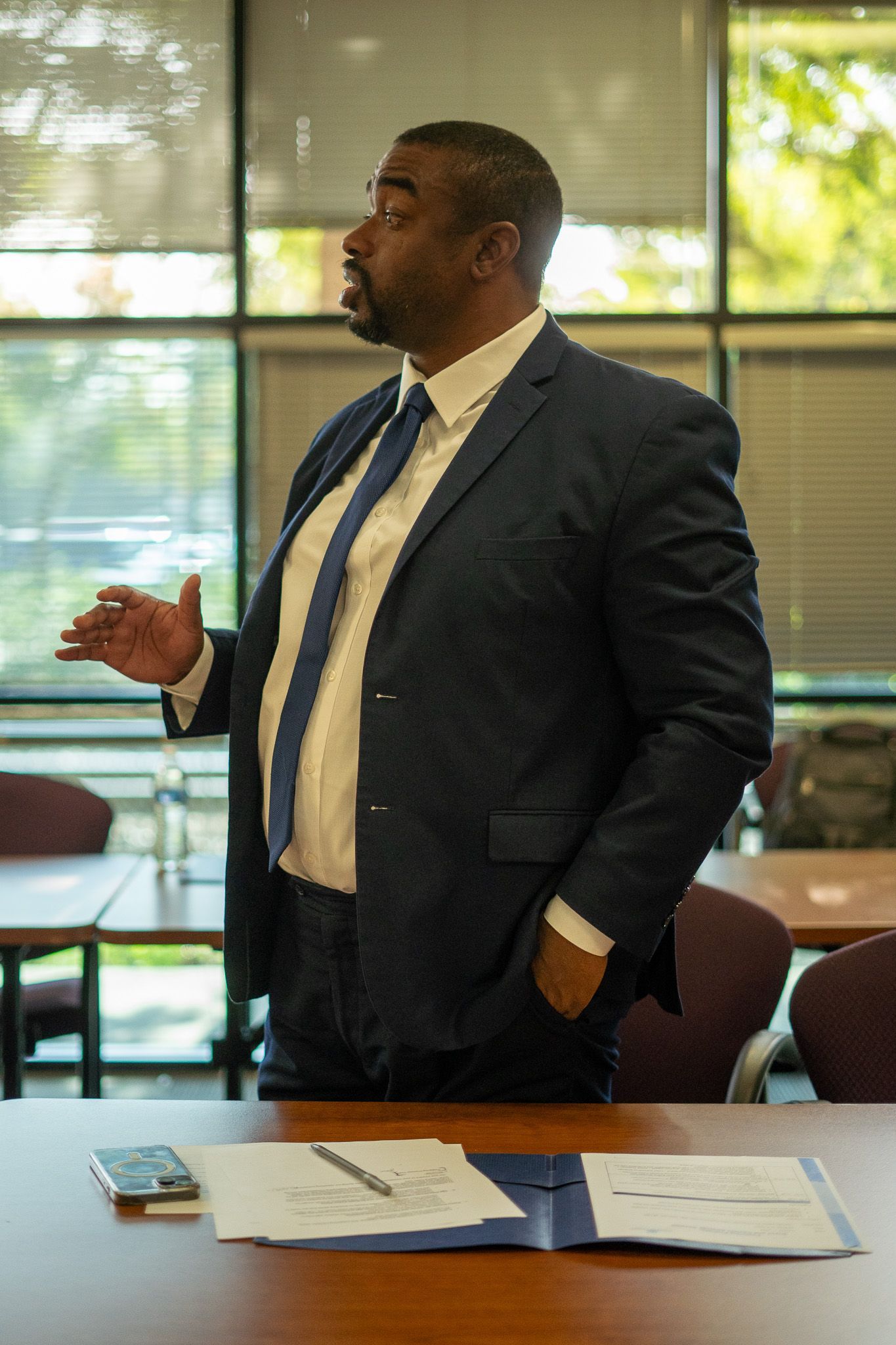 Man in a dark suit speaking in a conference room beside a table with papers and a phone