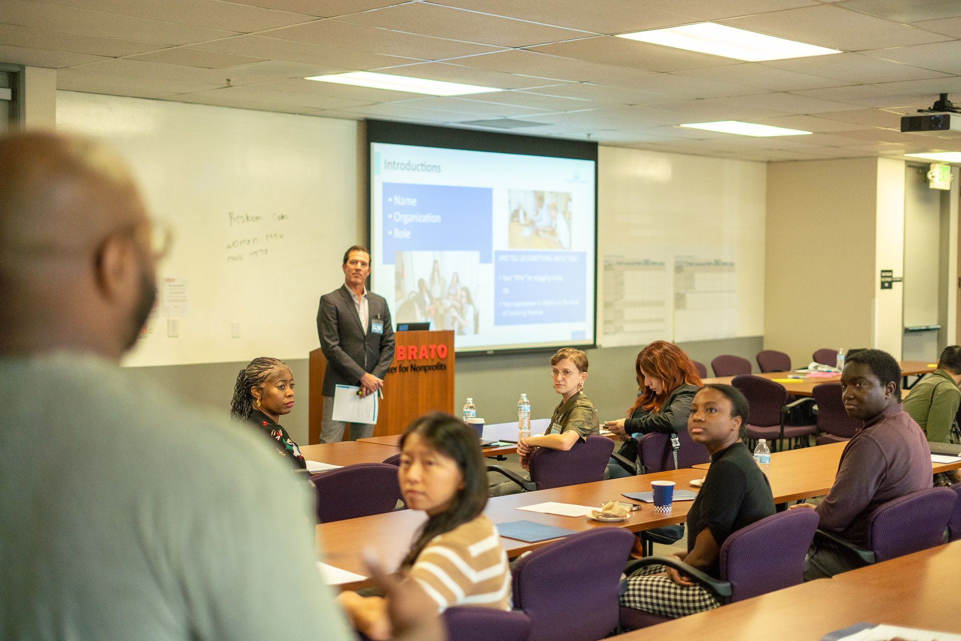 Classroom presentation with speaker at podium, projected slides, and seated audience at tables