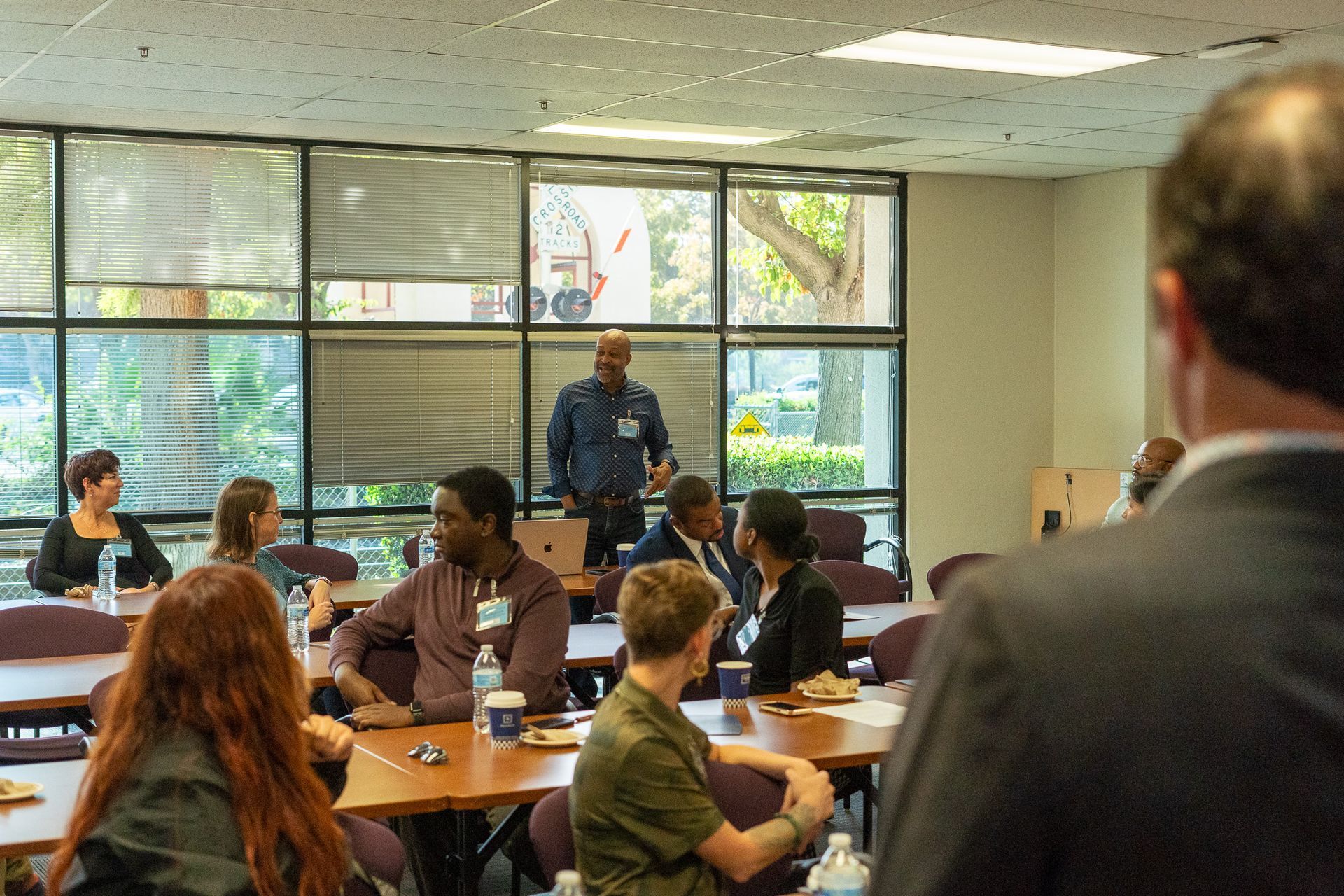 Meeting room with people seated at tables while a speaker stands at the front near large windows.