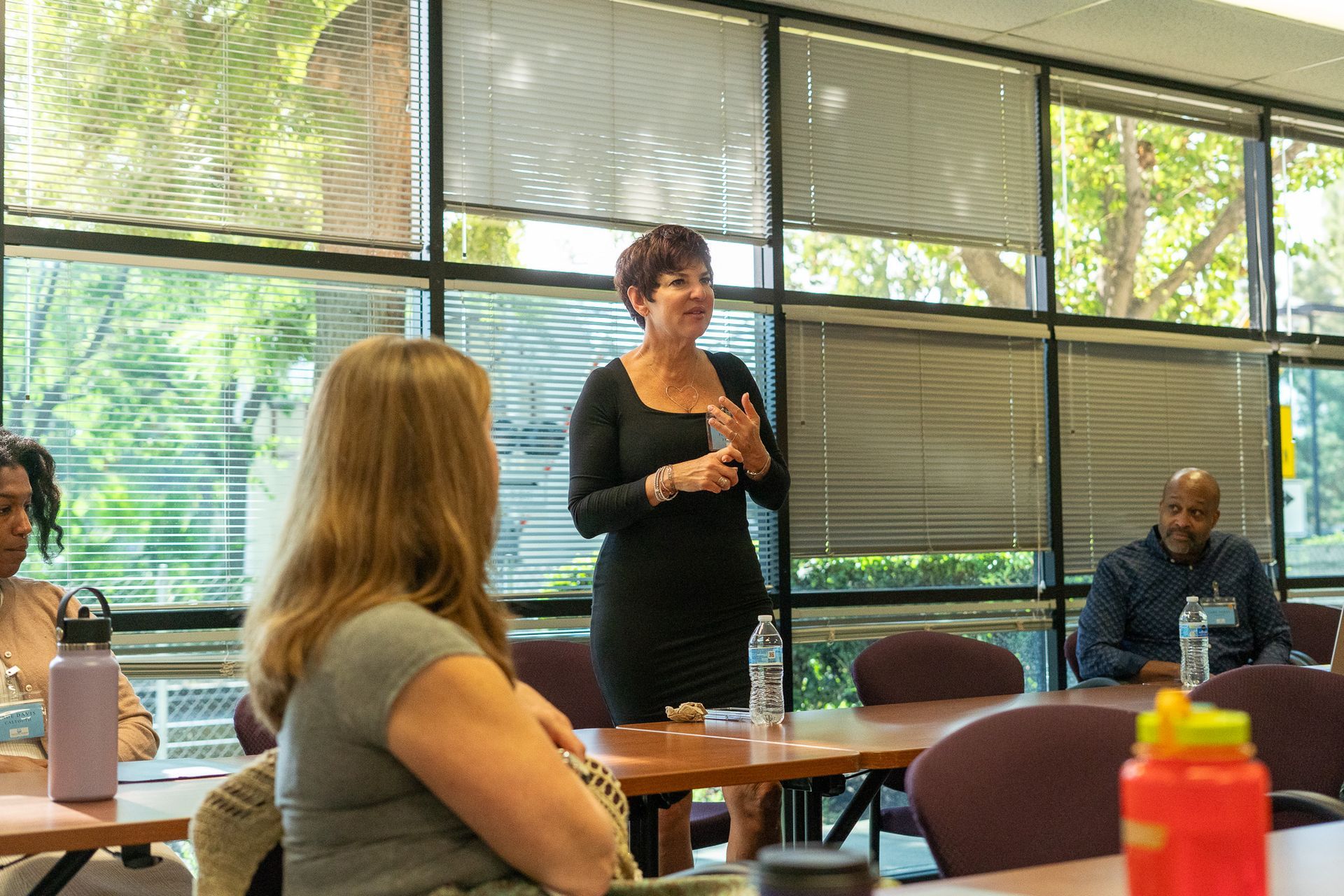 Woman speaking in a conference room while others listen at tables near large windows.
