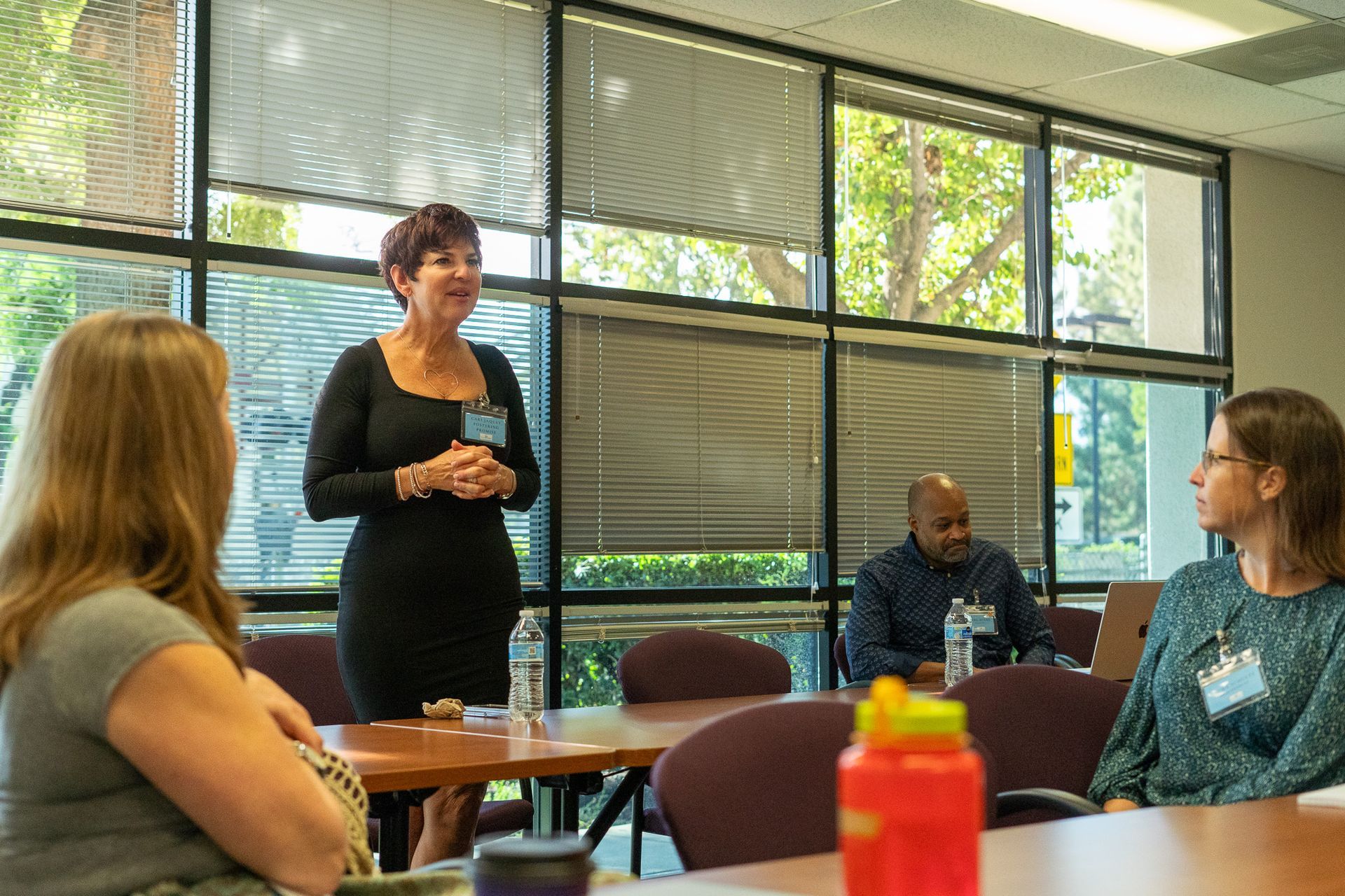 Small group meeting in a bright room, with one woman standing and speaking to seated attendees.