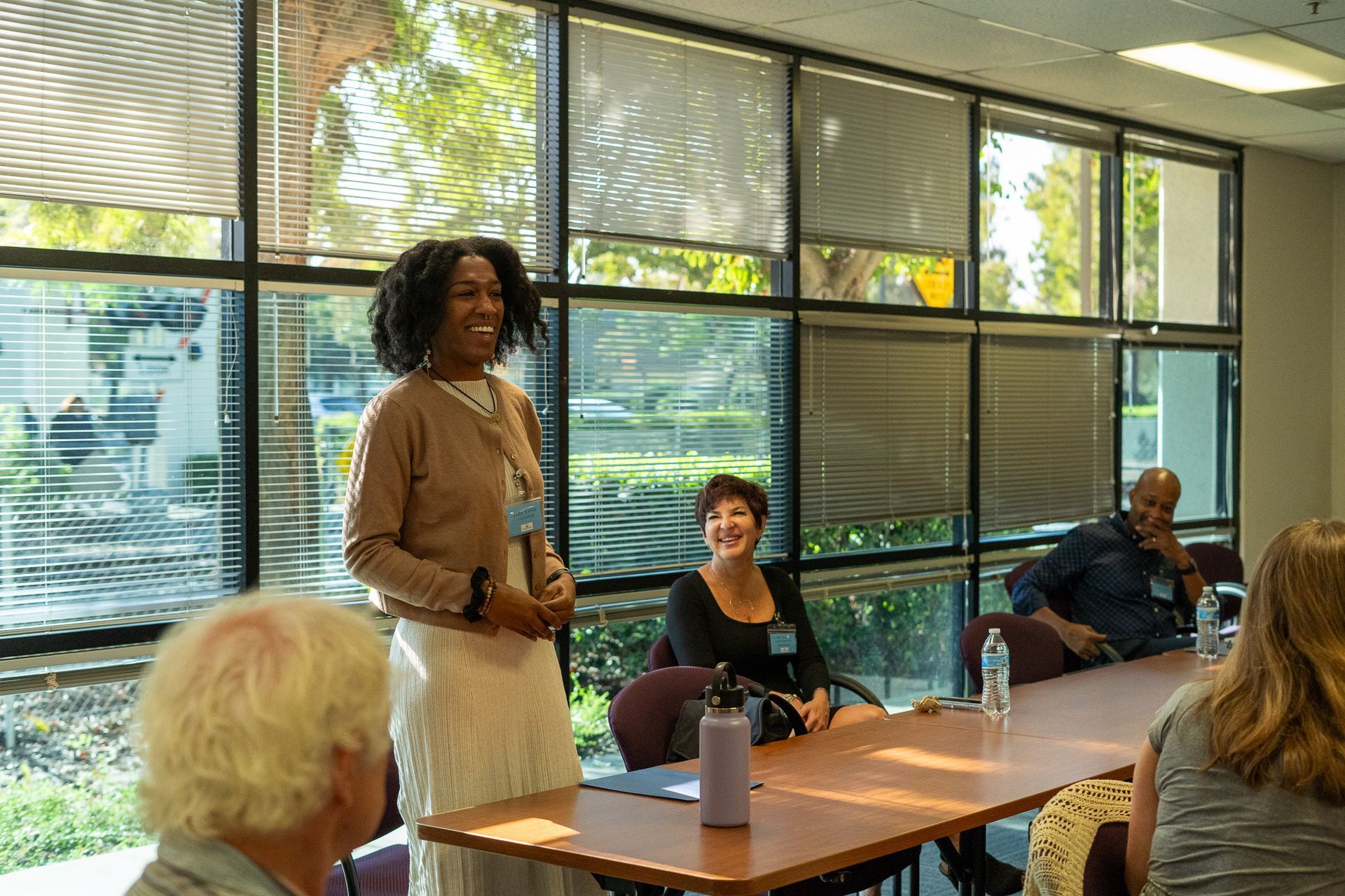 Speaker presenting in a conference room with seated audience and large windows behind them.