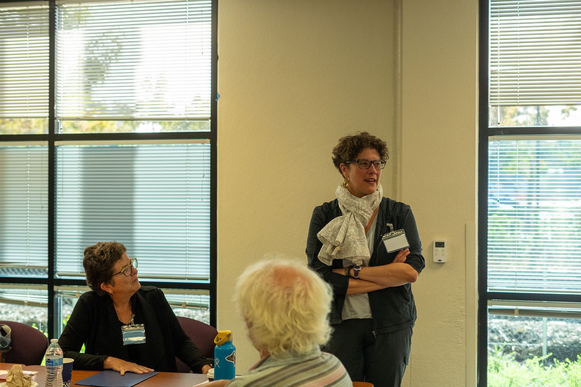 Person speaking to a seated group in a sunlit room with large windows and blinds