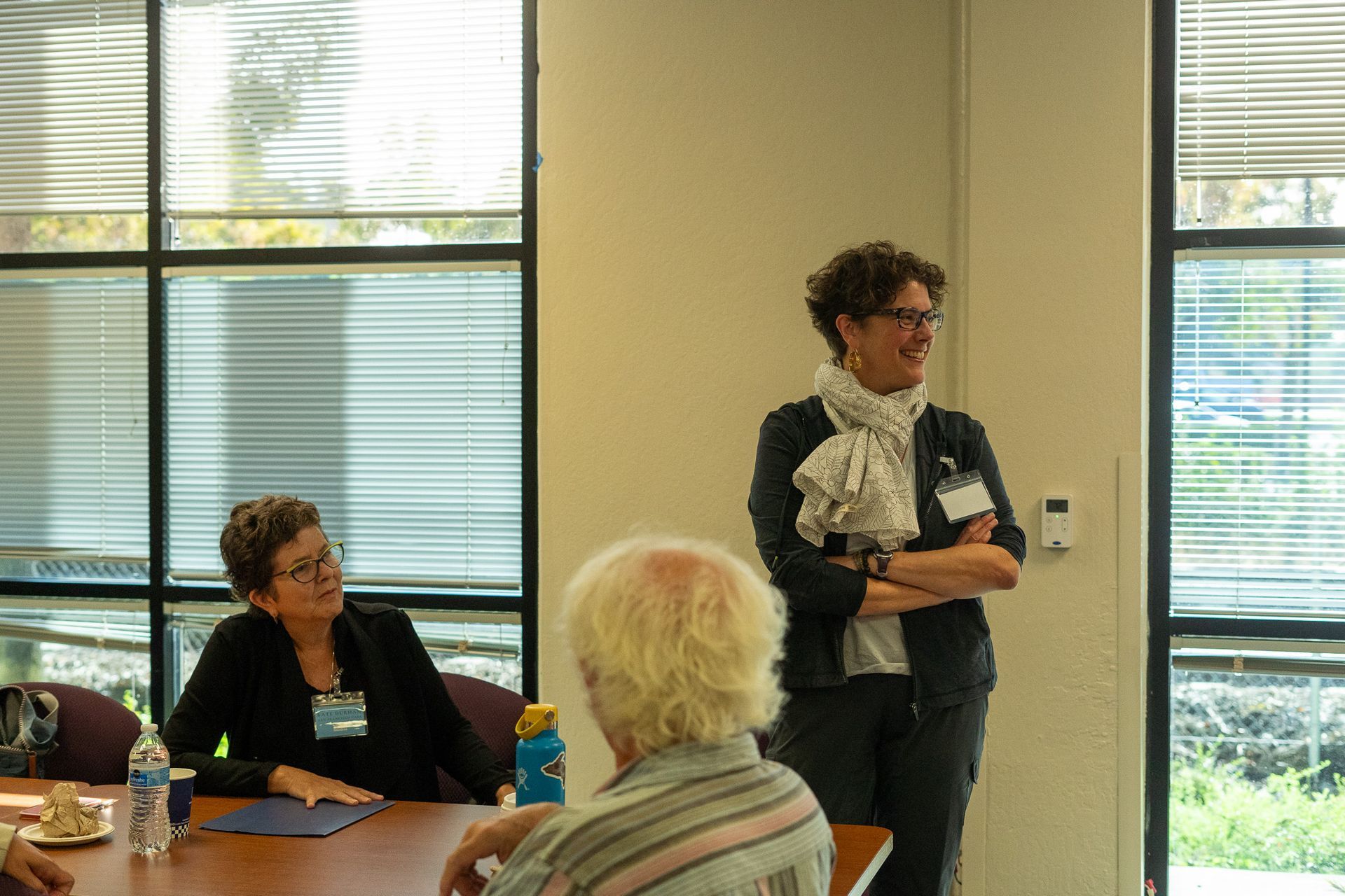 Two people speaking in a meeting room, with others seated nearby by large windows.