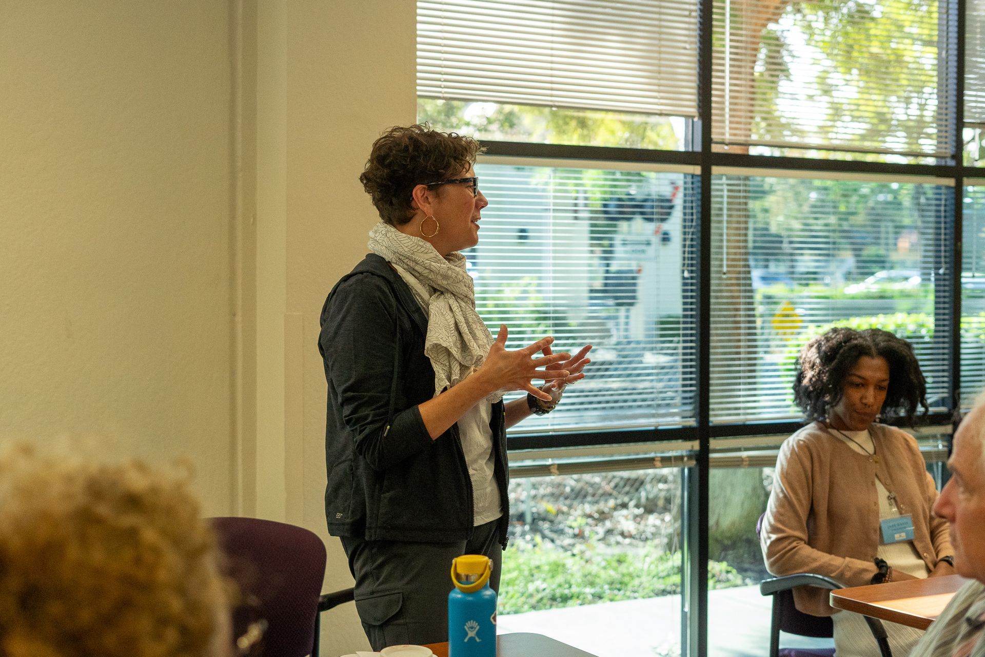 Person speaking in a meeting room beside a window, with others seated nearby.
