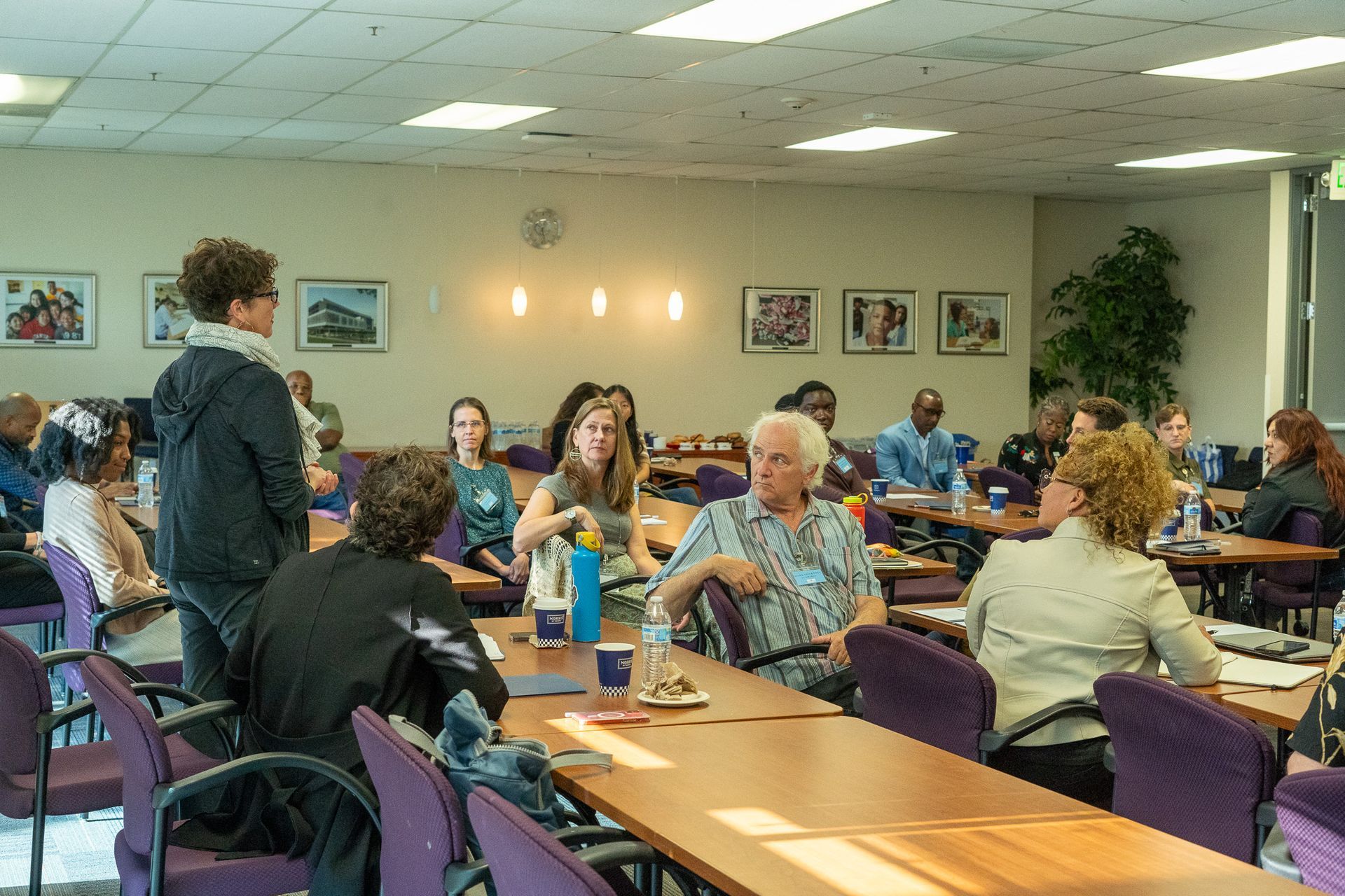 People seated at tables in a community dining hall, talking and eating under fluorescent lights.