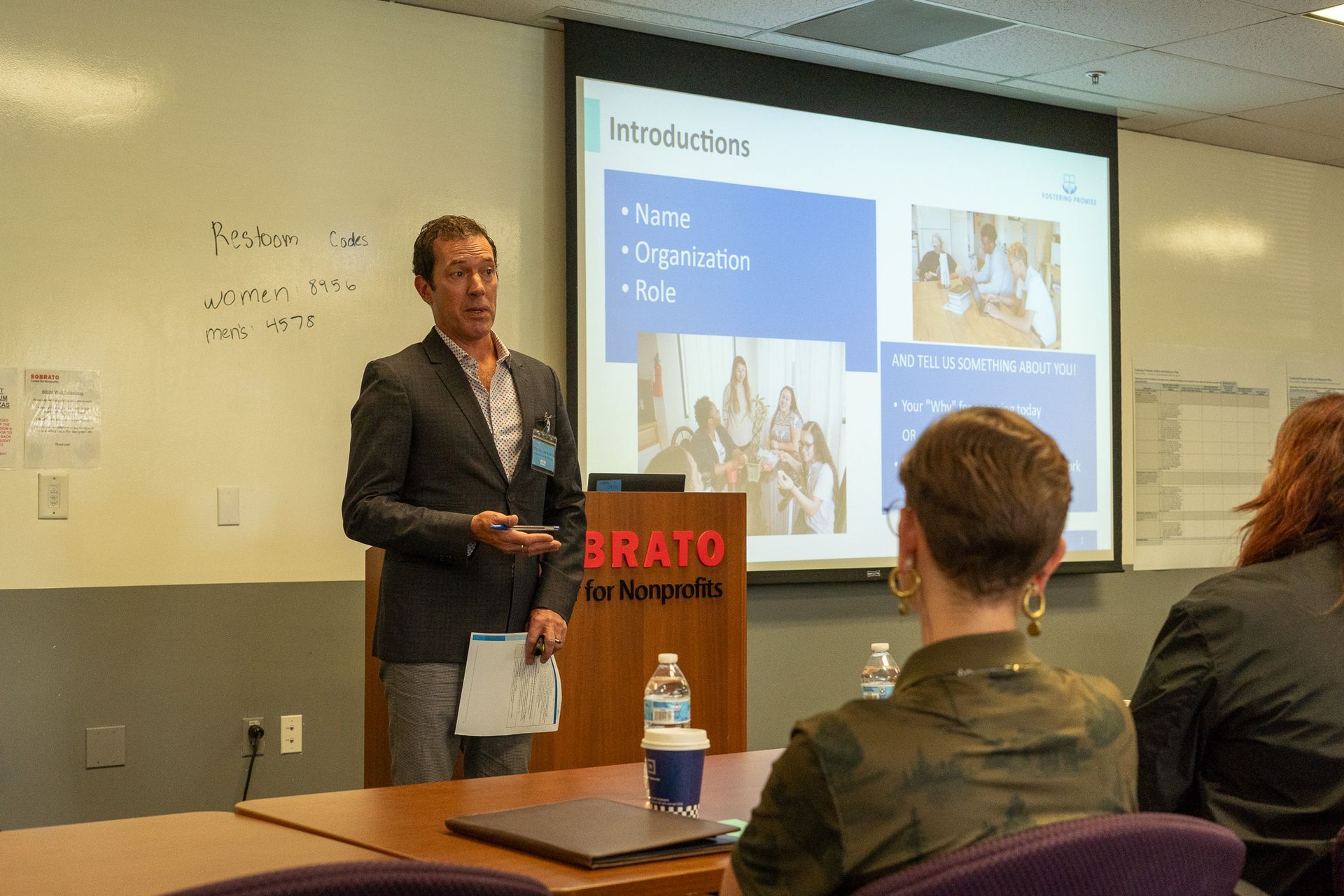 Presenter speaking at a podium during a classroom lecture, with a projection screen and seated audience.