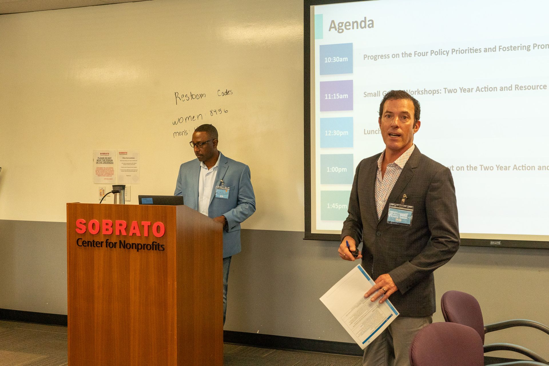 Two presenters at a conference podium and screen in a meeting room