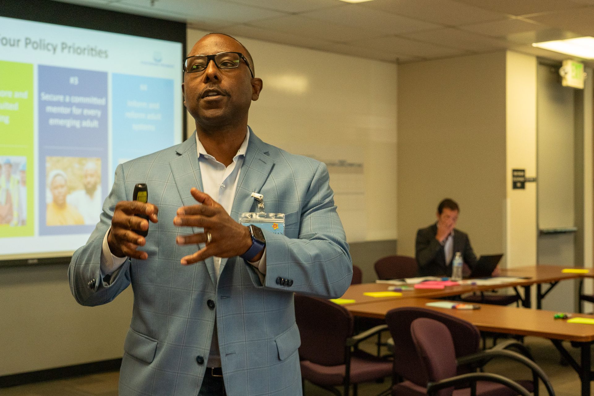Man speaking in a classroom, gesturing in a light blue blazer, with a presentation slide behind him.