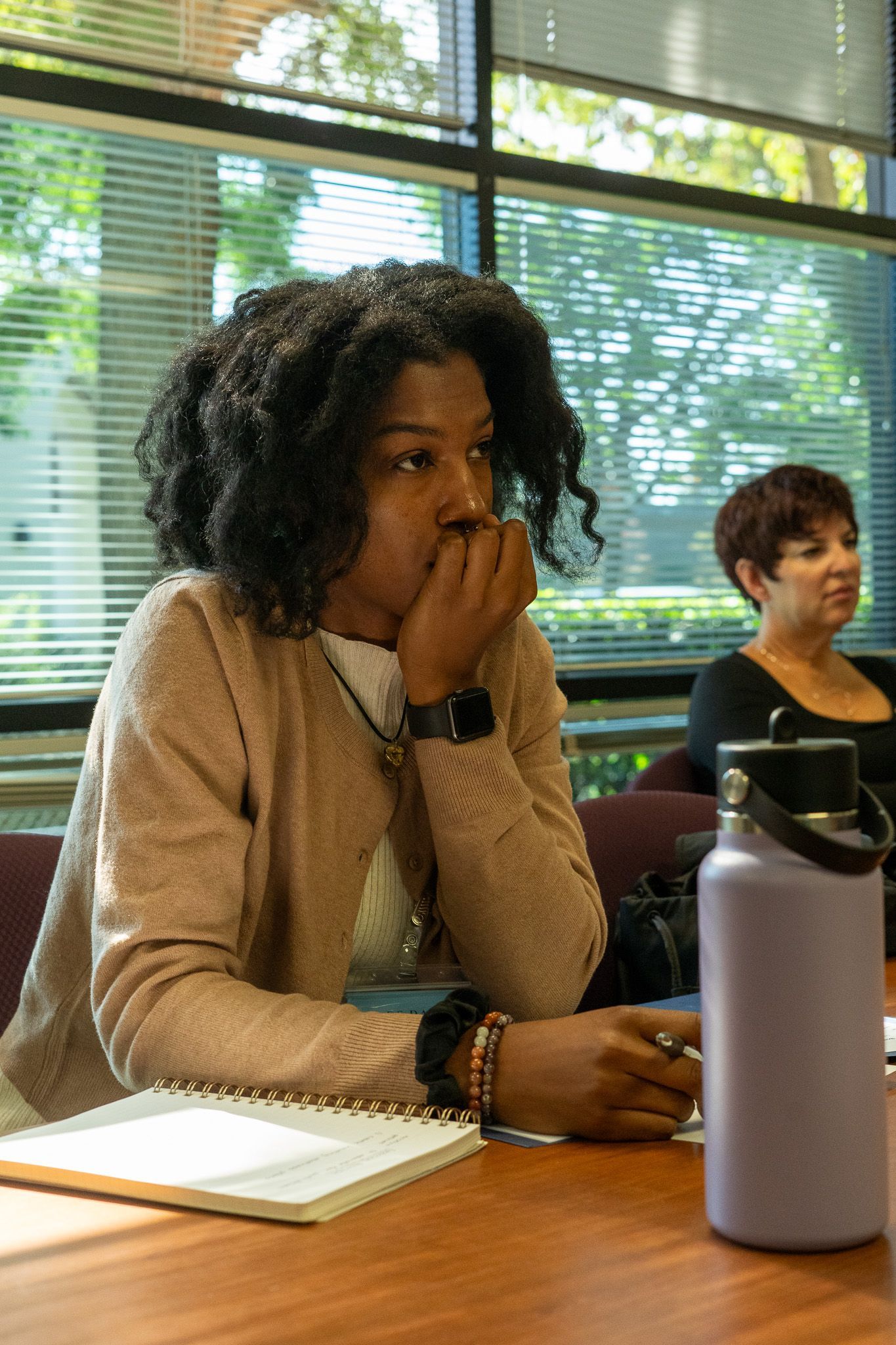 People sitting at a table in a bright classroom, one with hand over mouth, notebook and water bottle in front