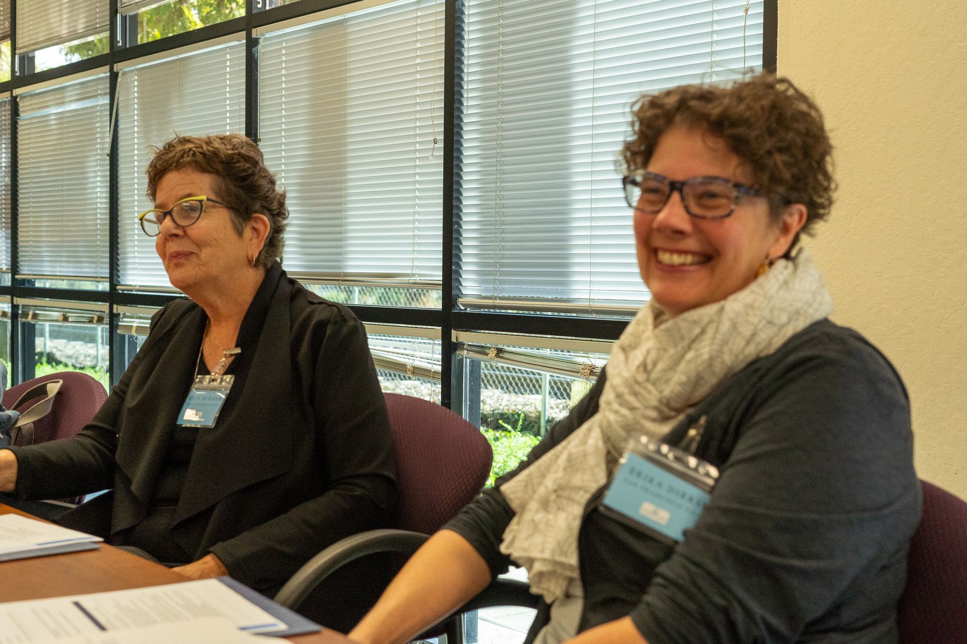 Two people seated at a table in a bright meeting room, smiling and wearing name tags.