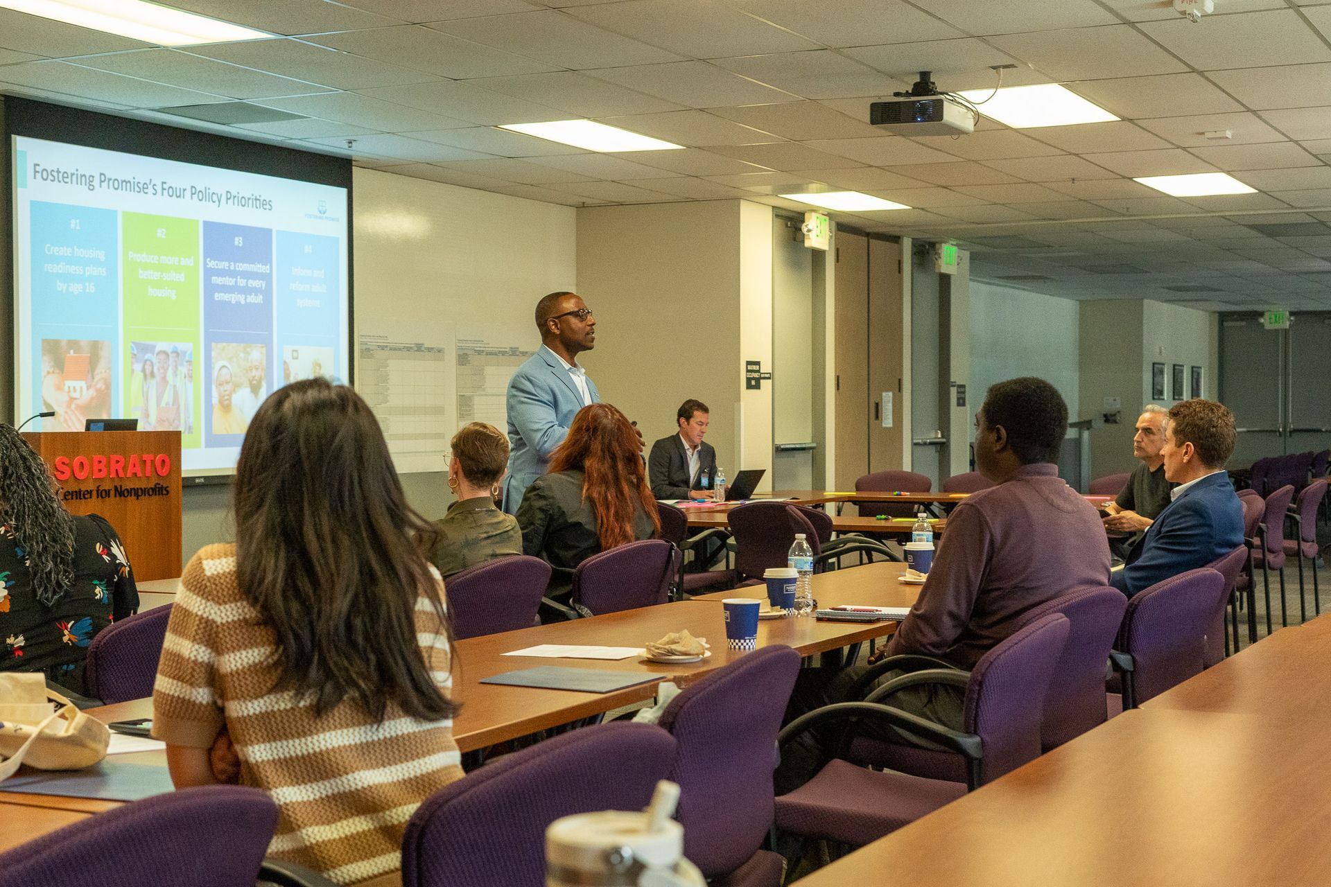 College classroom with presenter at projector screen and students seated at desks, discussing a presentation