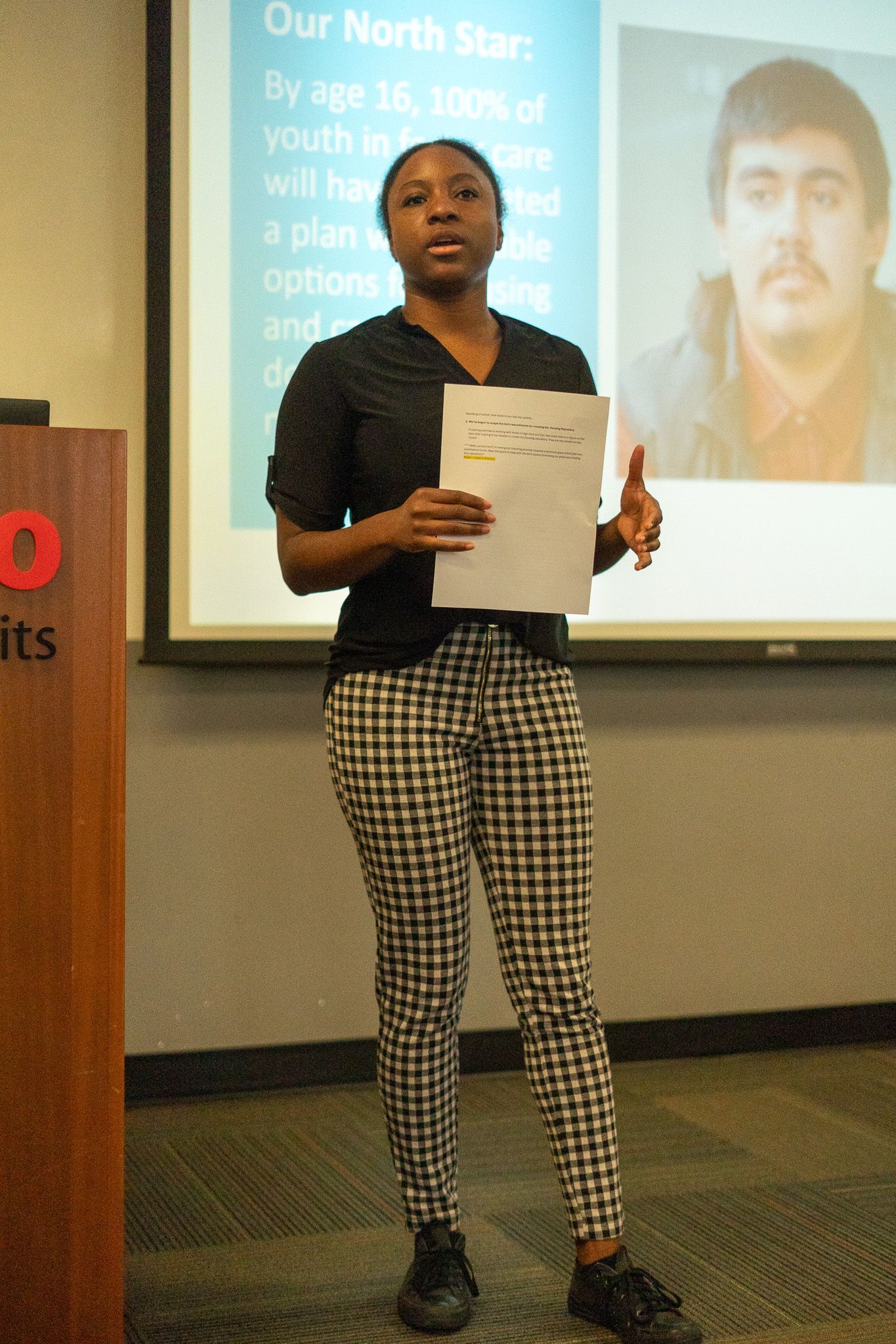 Woman presenting a paper in a classroom, standing before a projected slide and portrait photo.