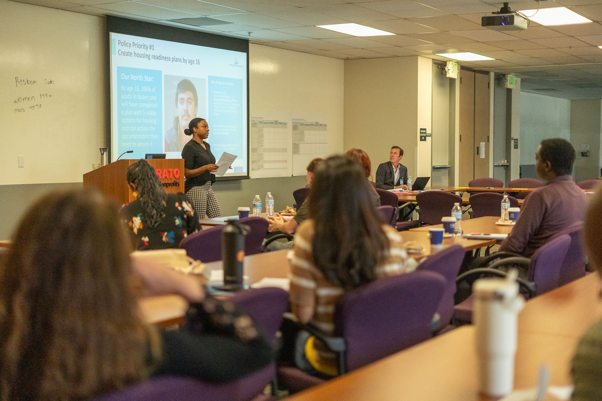 Classroom lecture with presenter at front and audience seated at desks watching presentation