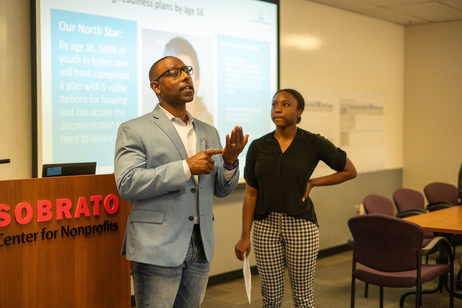 Two people presenting in a classroom, with a projected slide and tables and chairs in the background.