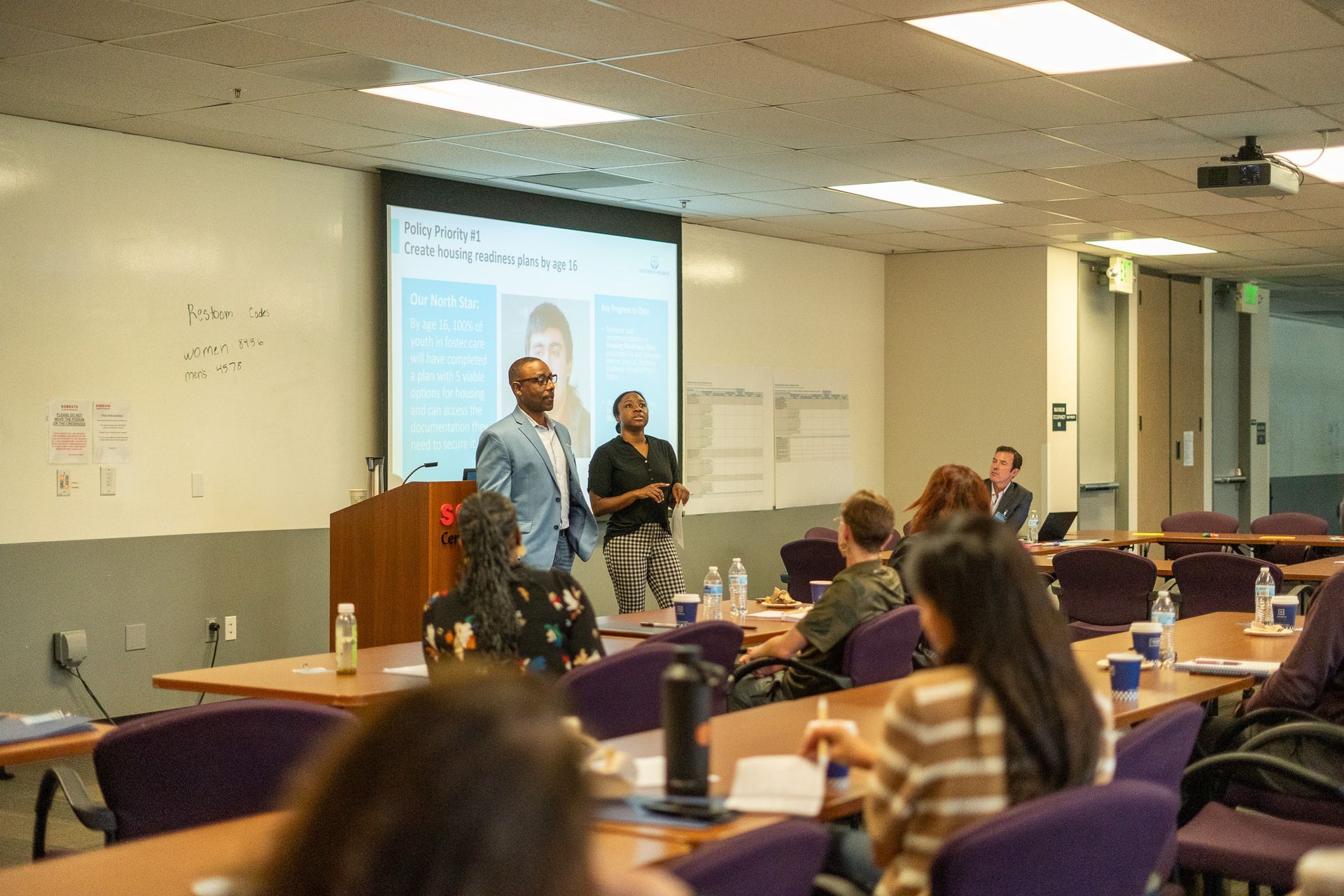 Presentation in a classroom with two speakers near a projector screen and students seated at desks
