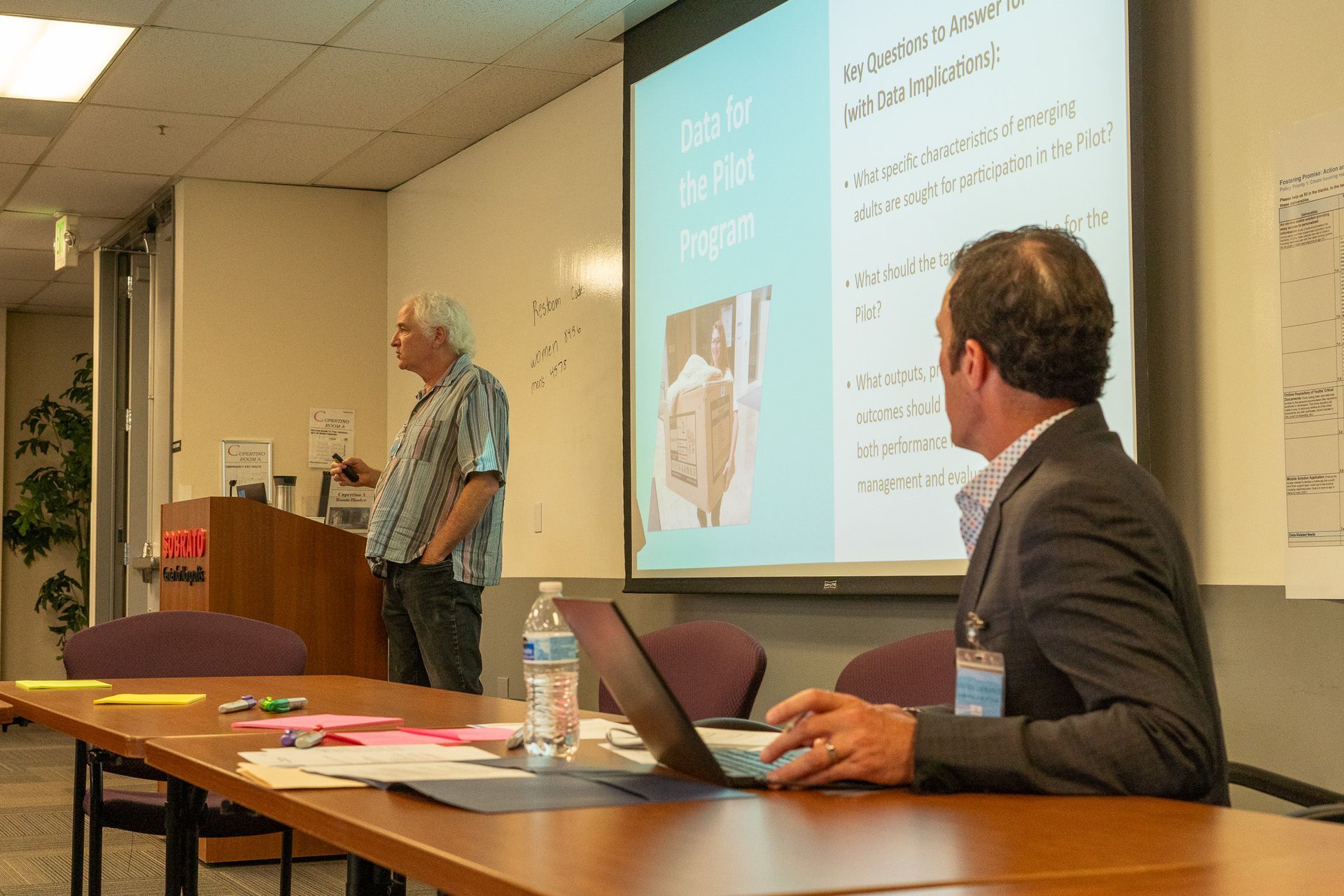 Presenter speaking in classroom while seated man listens at a laptop, with projected slides nearby