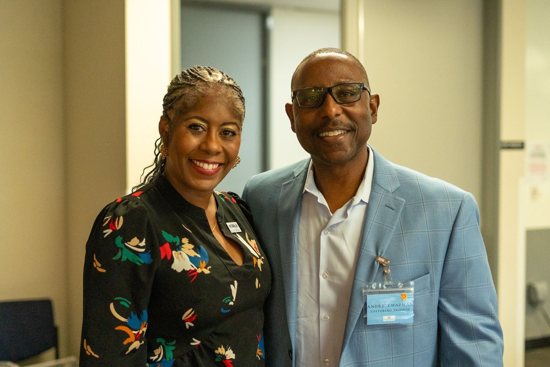 Two people smiling indoors, standing close together; one wears a black floral top, the other a light blue blazer.